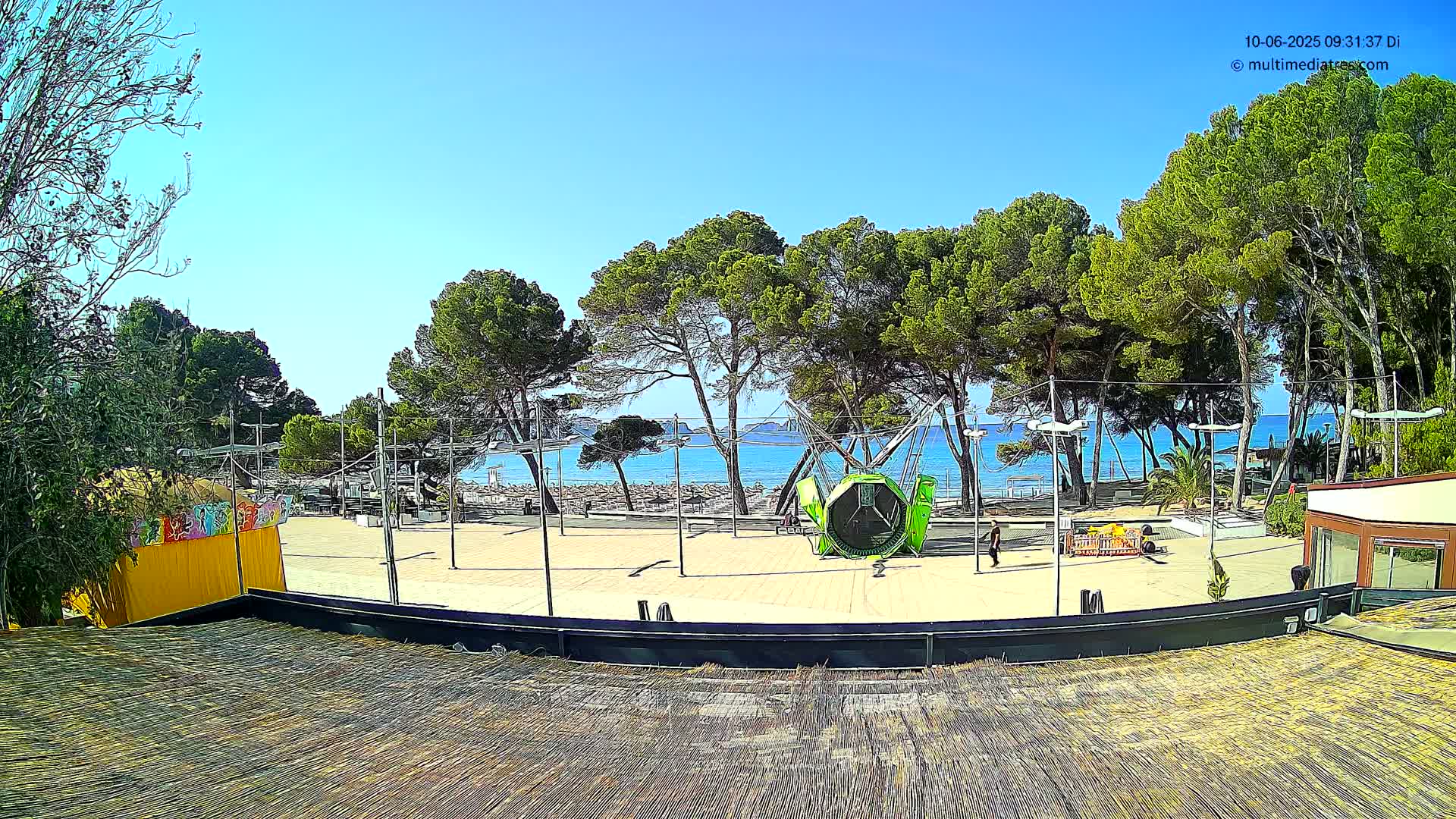 A beachfront plaza with a playground, surrounded by pine trees under a clear blue sky.