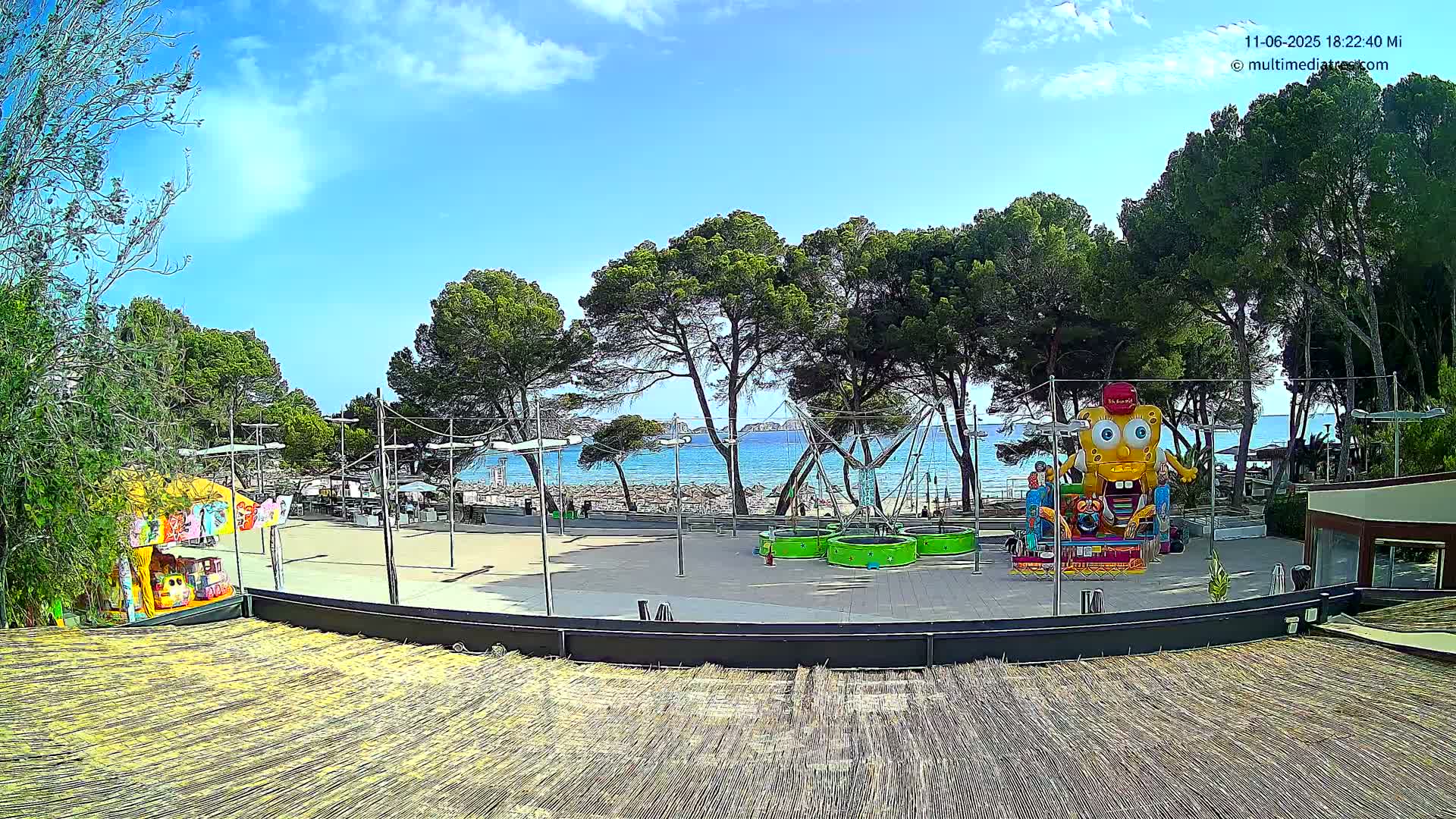 A beachfront amusement area with several rides and a trampoline, under a mostly sunny sky with a few clouds.