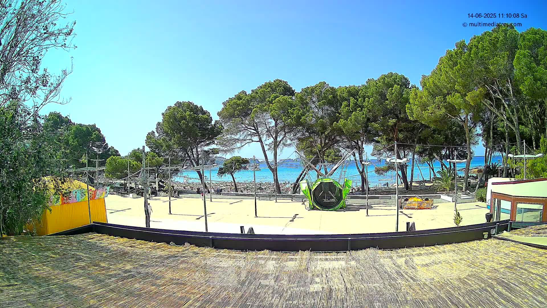 A sunny outdoor scene shows a paved area with a playground structure near a beach, behind a line of pine trees.