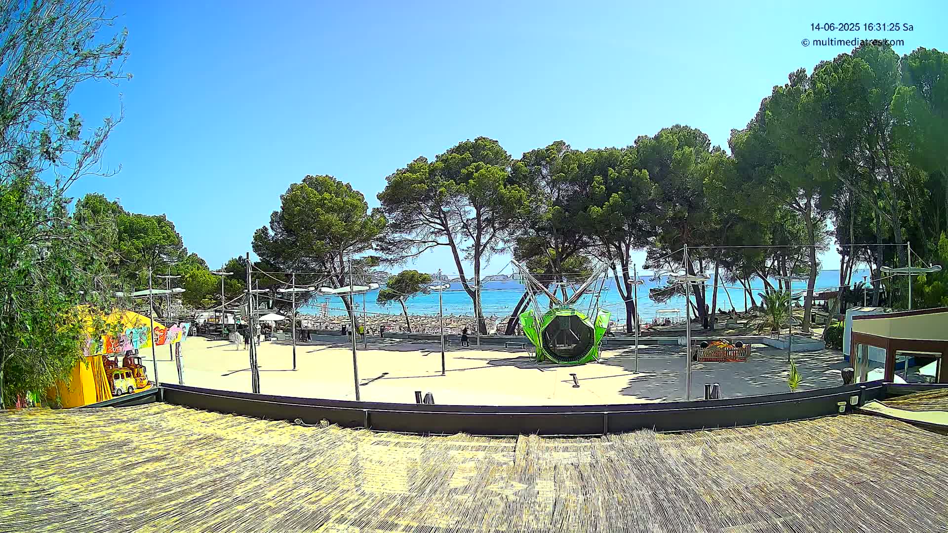 A sandy area with playground equipment is situated between a line of pine trees and a sunny beach with clear turquoise water.