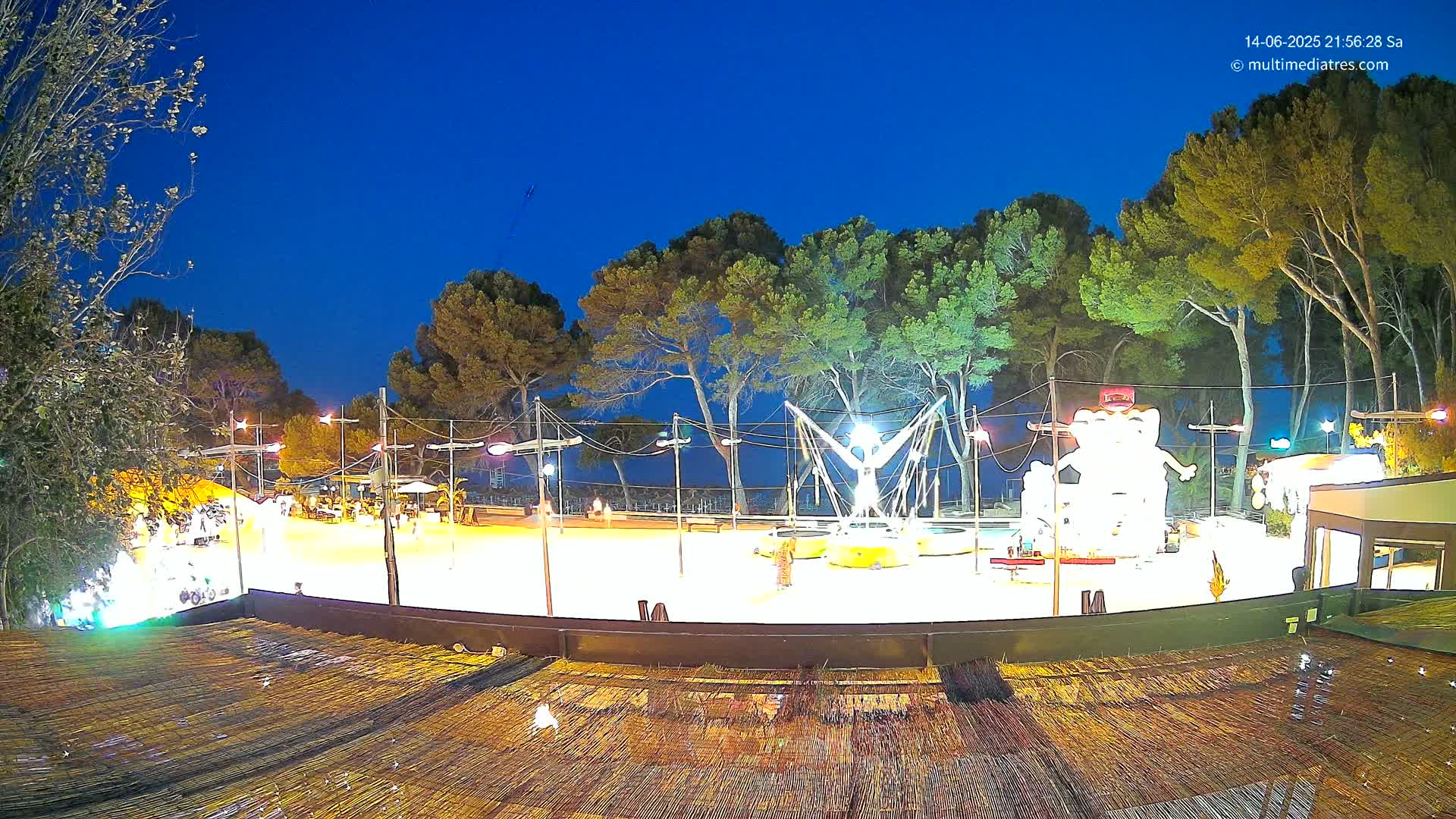 A sandy area with playground equipment is situated between a line of pine trees and a sunny beach with clear turquoise water.