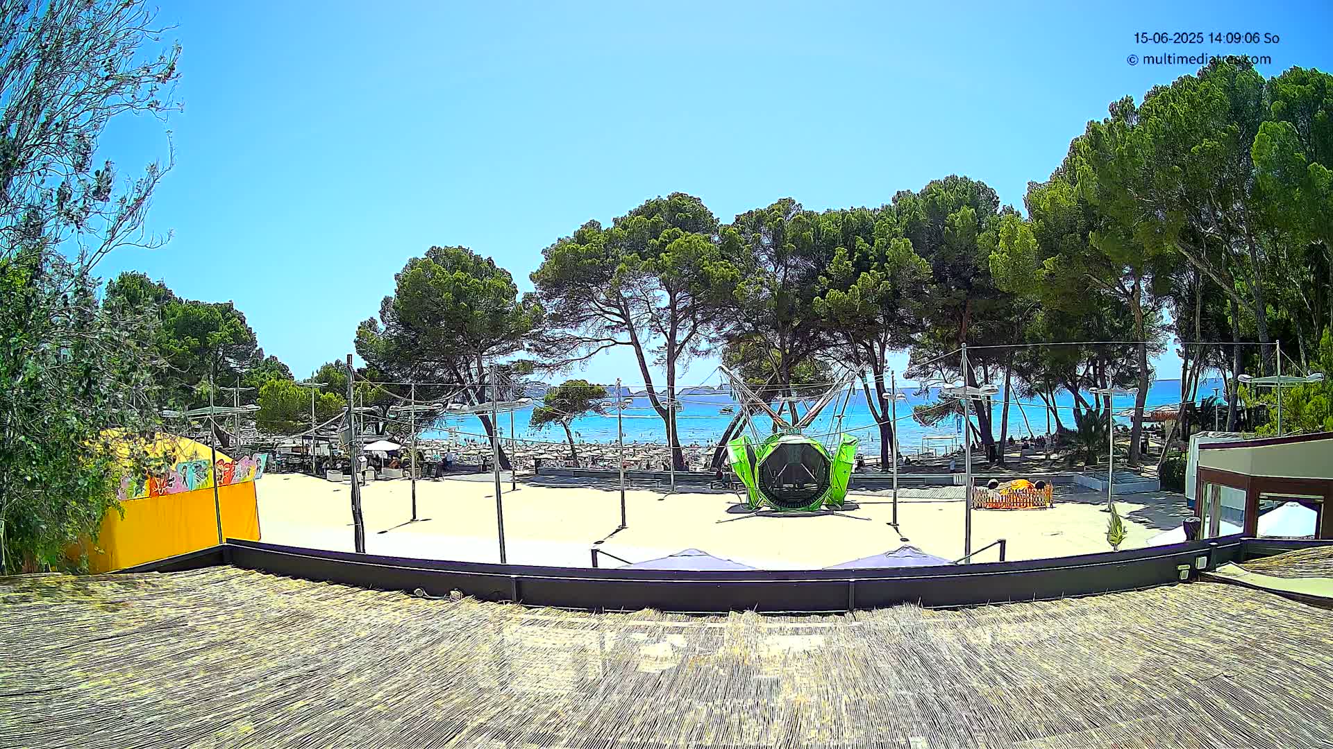 A sandy beach area with a green playground ride is visible beyond a line of pine trees under a clear blue sky.