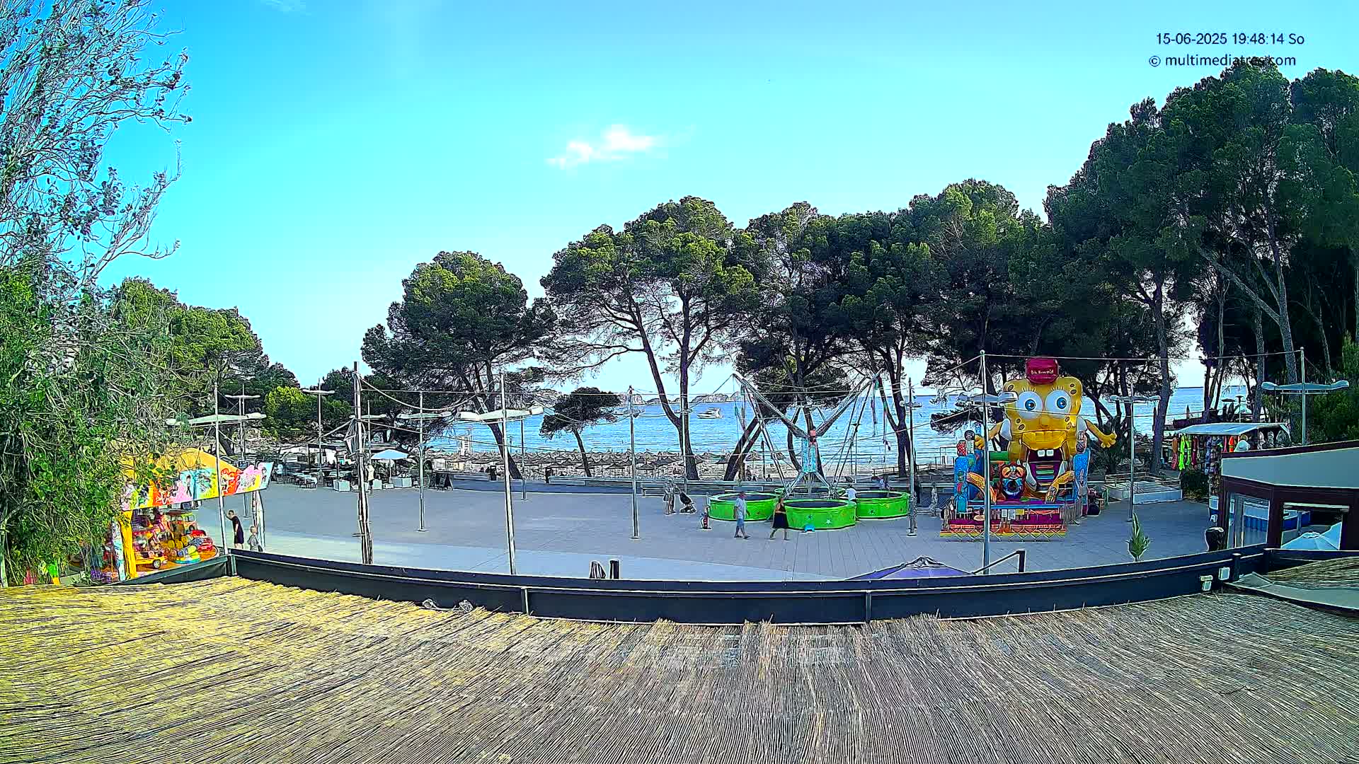 A seaside plaza with a bouncy castle, small kiosks, and trees under a clear blue sky.