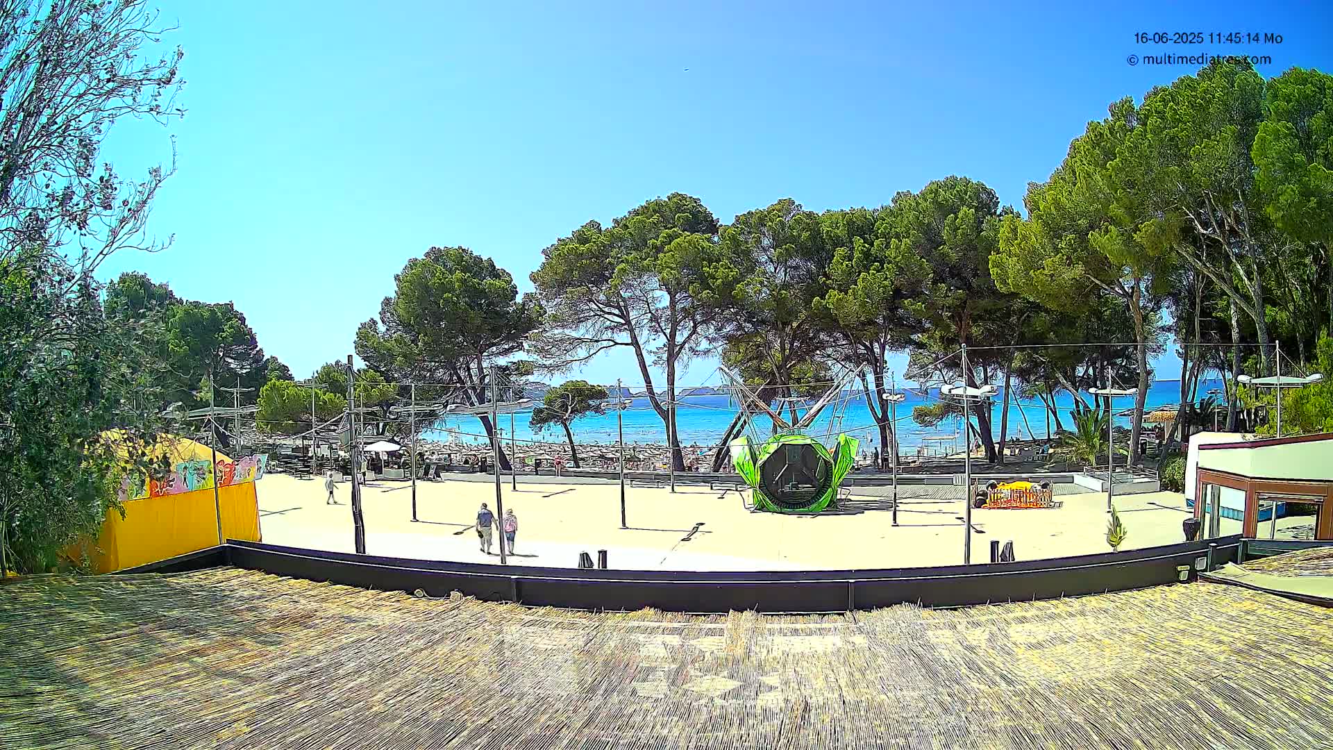 A sandy beach area with a few people, some trees, and a beachside playground is visible under a clear, sunny sky.