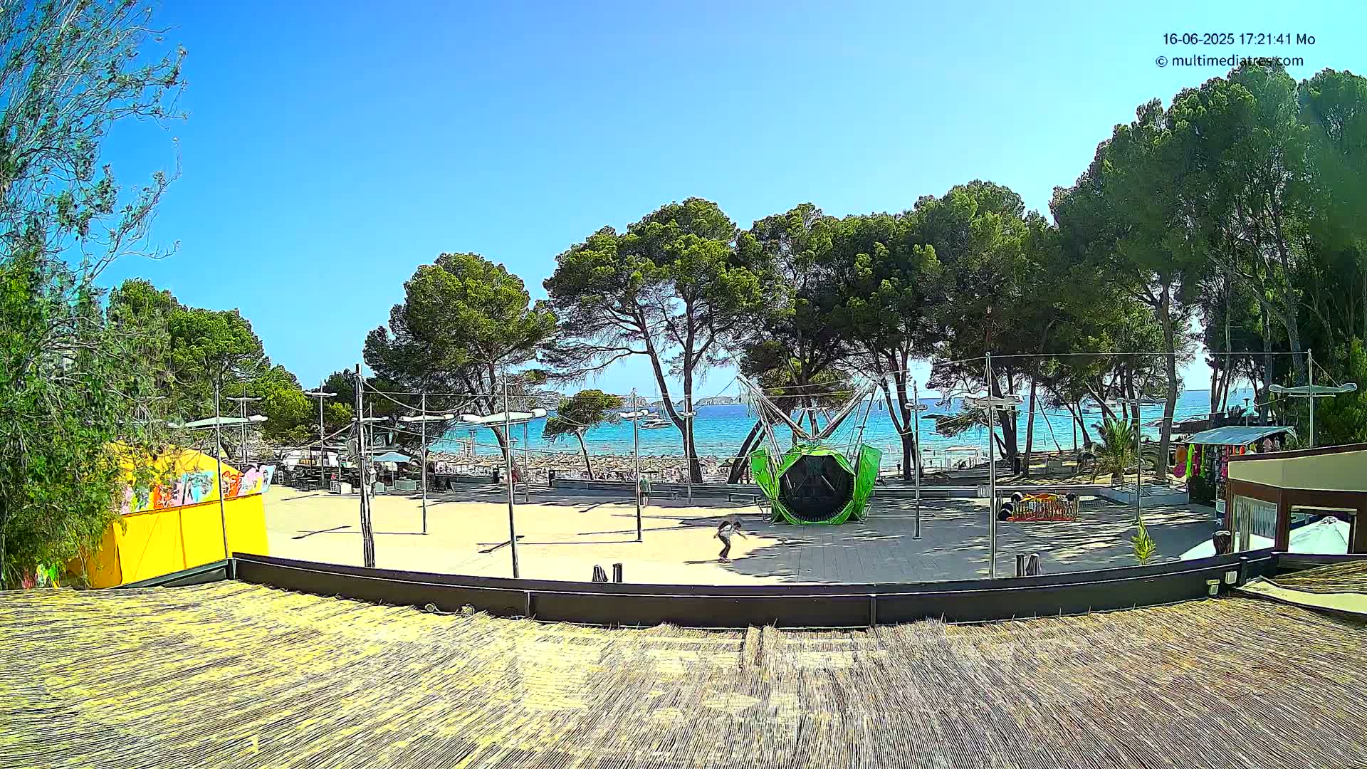 A sandy area with a playground and small shops is situated between a line of pine trees and a calm, blue ocean on a sunny day.