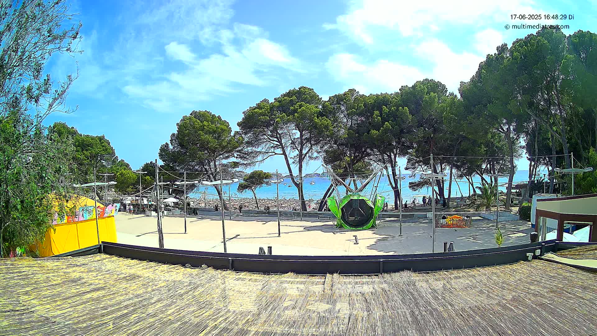 A sunny day at a beachside plaza features a green playground ride, surrounded by trees, with a sandy beach and calm blue ocean visible in the background.