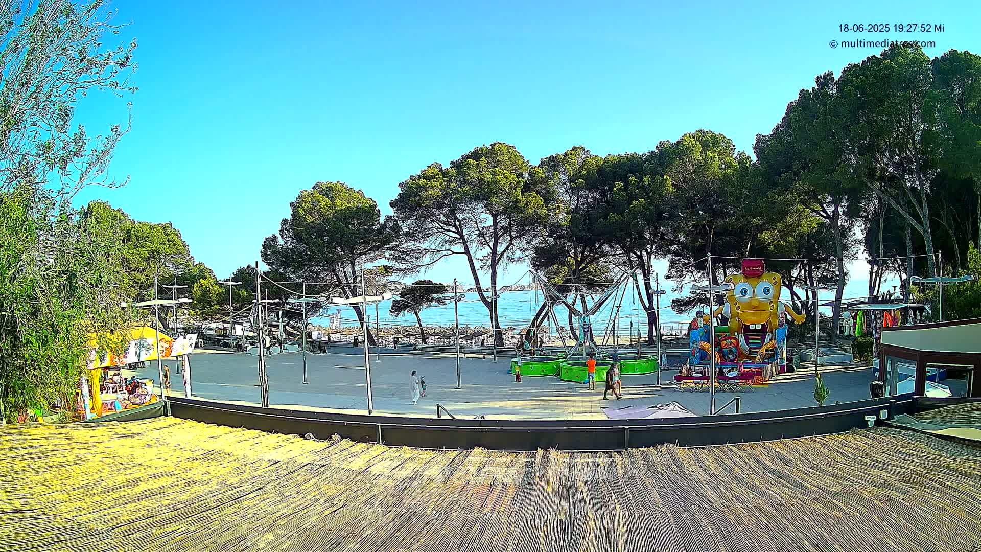 A seaside amusement area with a large inflatable character, playground equipment, and several trees under a clear blue sky.