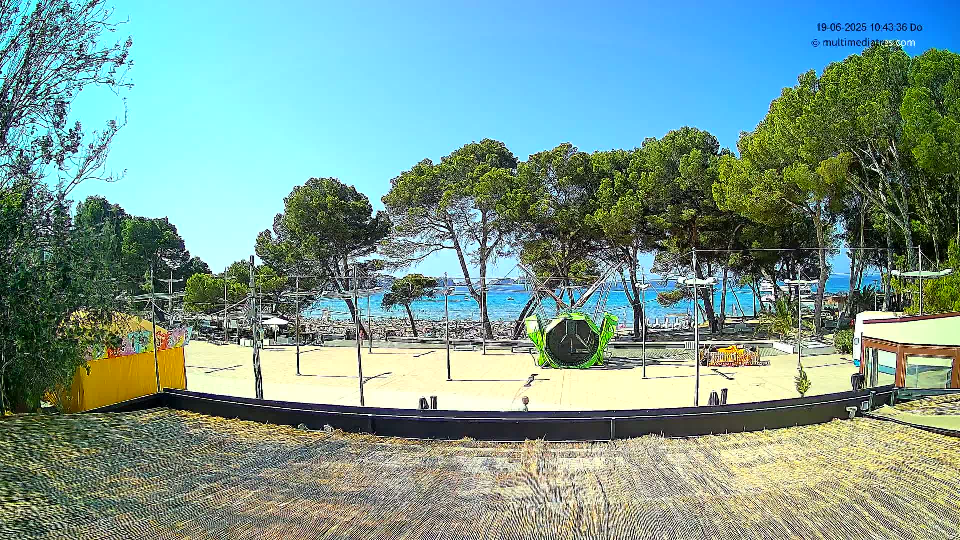 A sandy outdoor area with a playground and several trees overlooks a calm blue ocean on a sunny day.