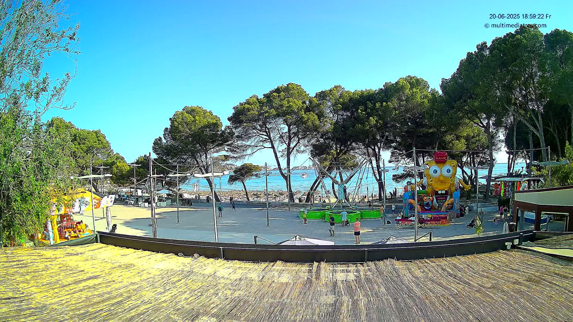 A sunny outdoor scene shows a paved area with amusement park rides near a beach, surrounded by pine trees.