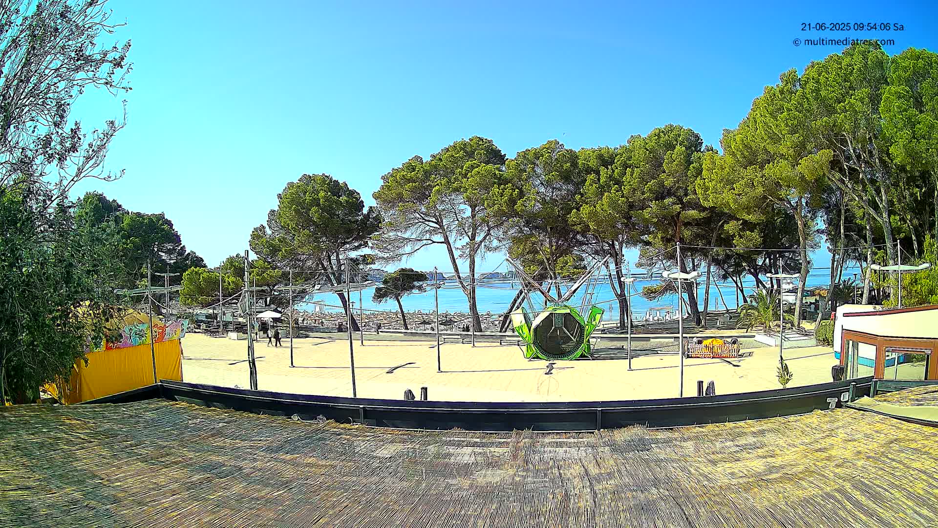 A sandy area with a green playground structure is situated near a beach, with a calm sea and many green trees under a clear blue sky.