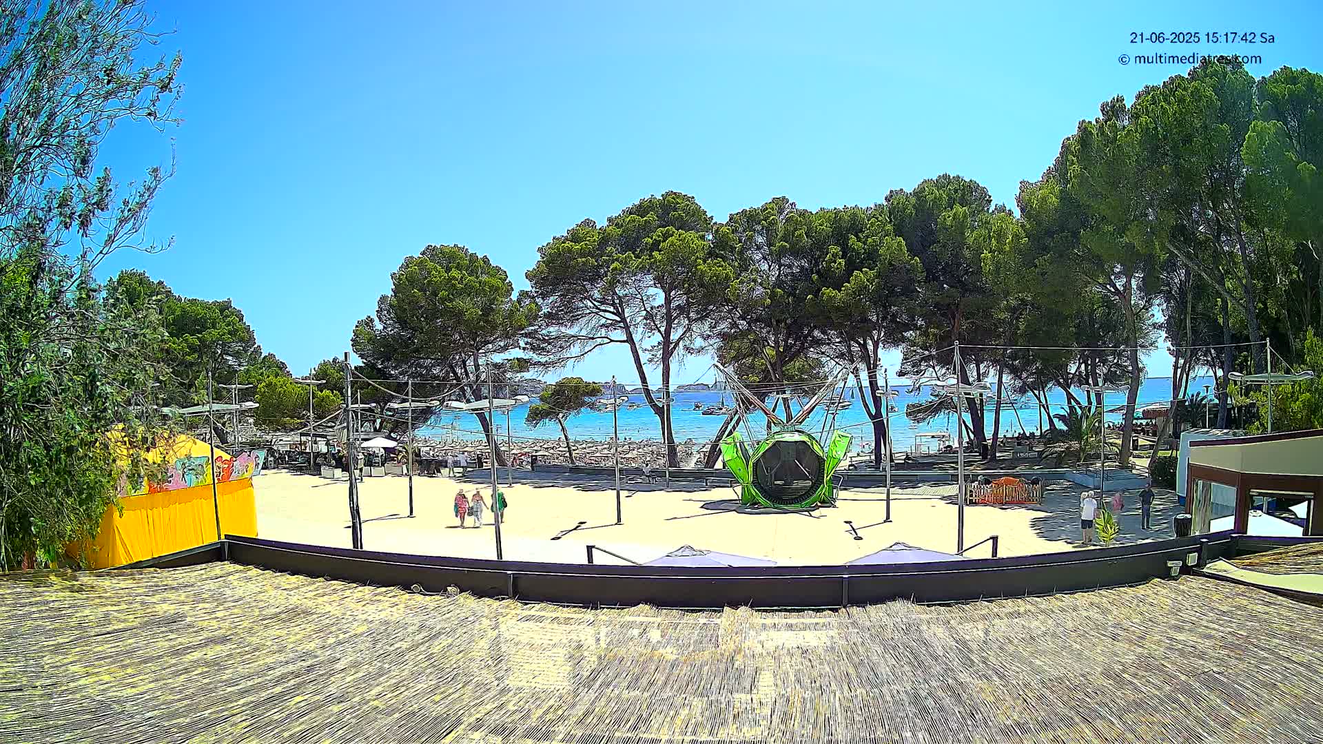 A sandy beach area with a playground and several people is visible beyond a row of pine trees under a clear, sunny sky.