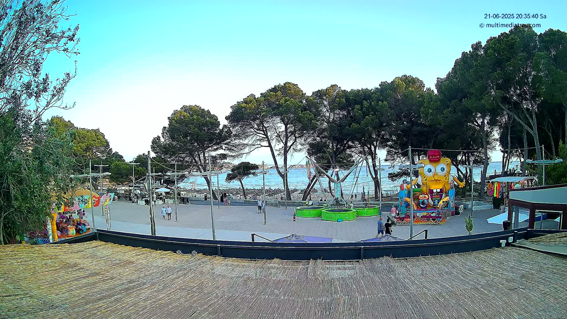An outdoor amusement area near a calm ocean, under a clear blue sky, features various rides and games amongst trees and people.