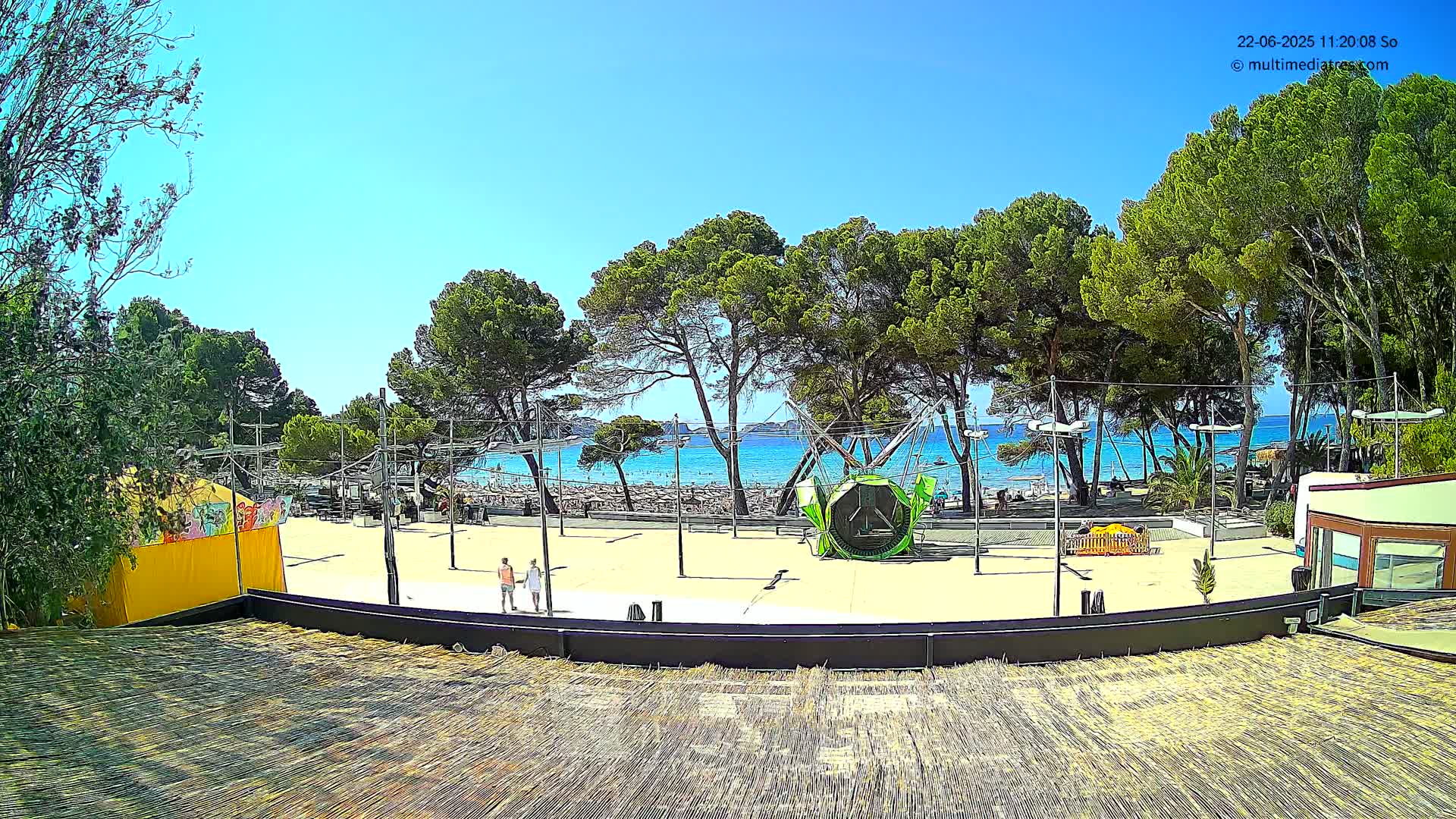 A sandy area with a playground and a few people, bordered by trees and overlooking a sunny beach and calm blue ocean.