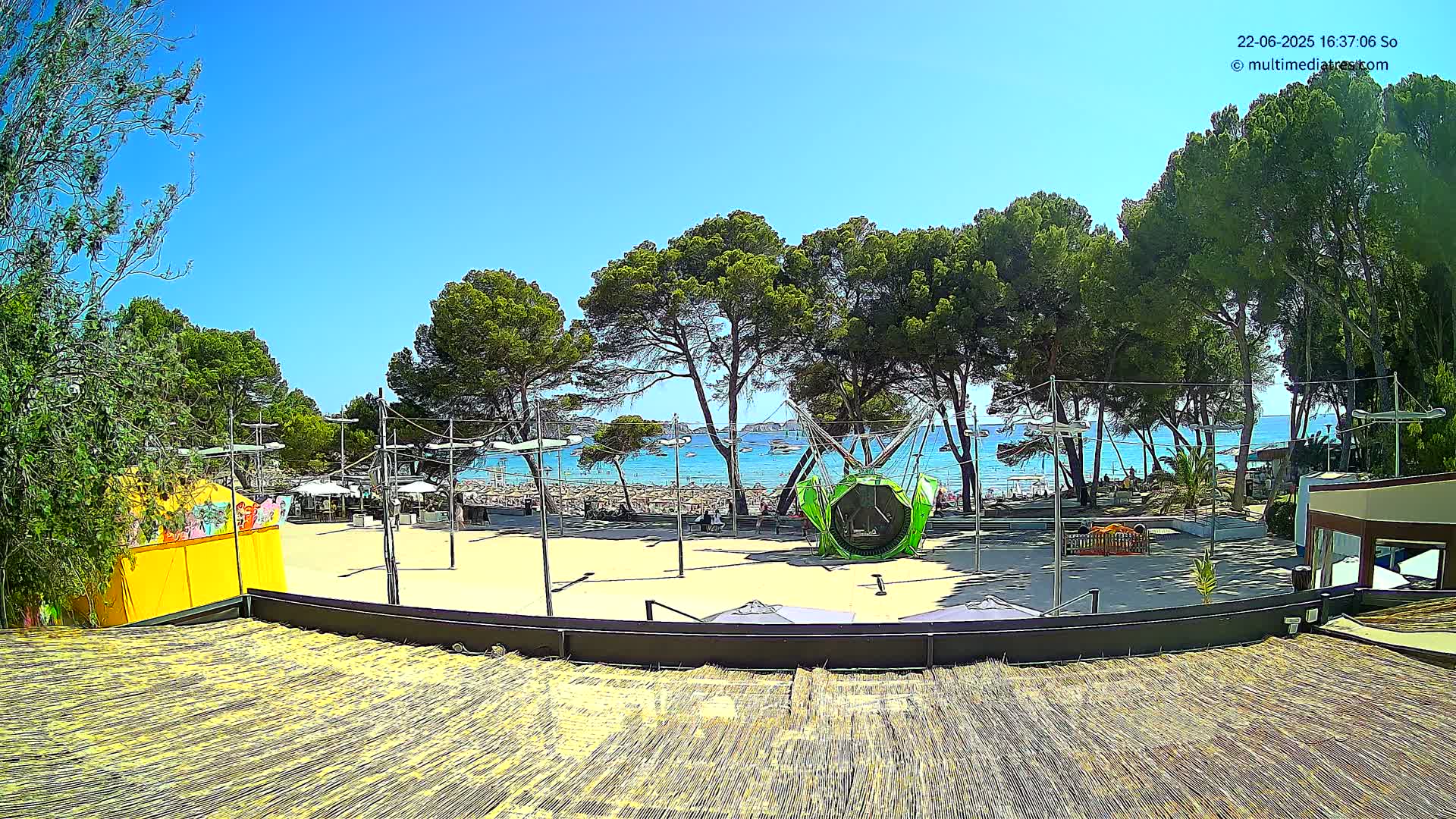 A sunny beach scene is viewed from an elevated position, showing a sandy play area with various play structures, surrounded by trees, with a calm blue sea and boats in the background.