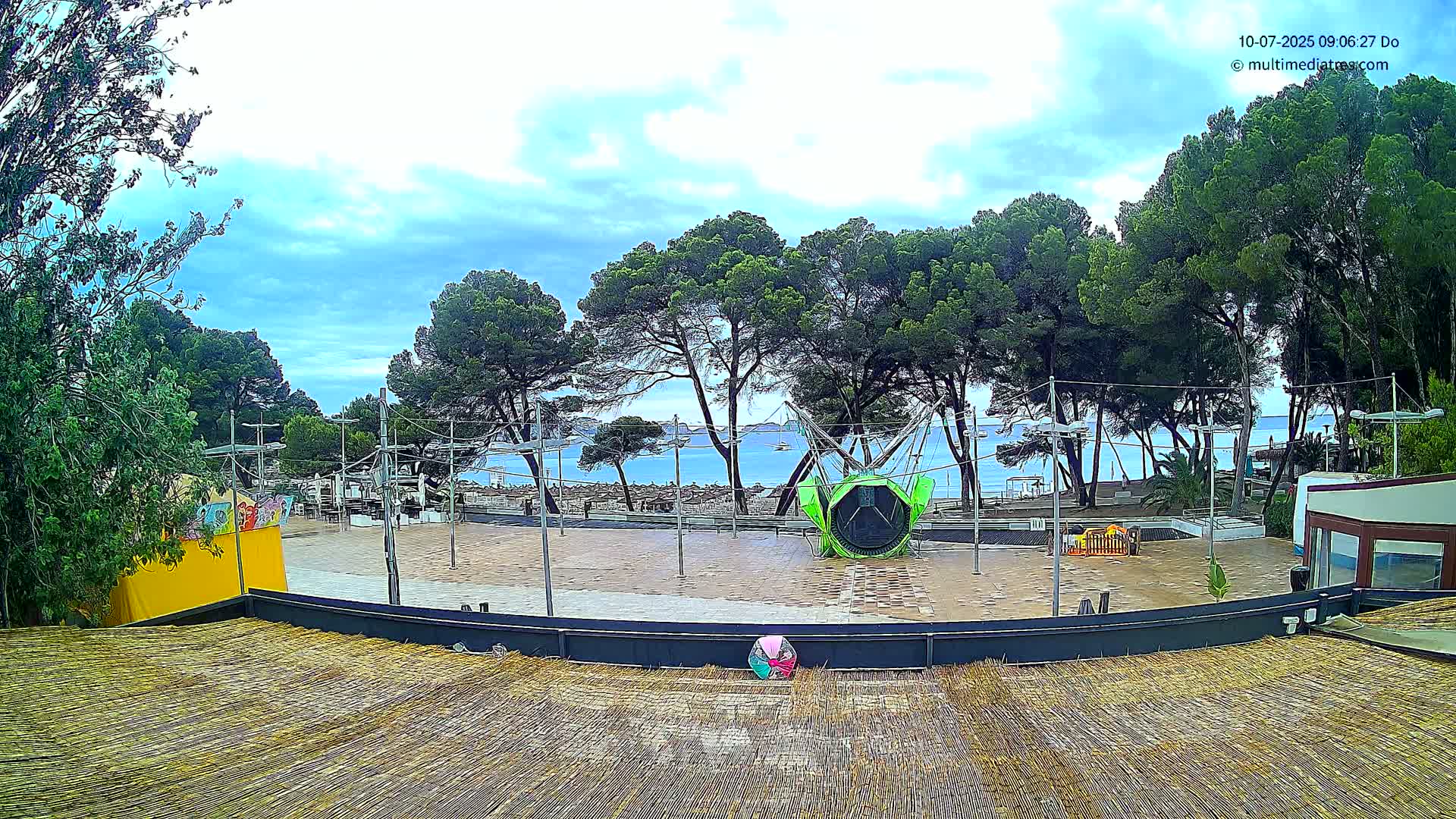 A partly cloudy day reveals a seaside plaza with a trampoline, surrounded by pine trees and a view of calm water.