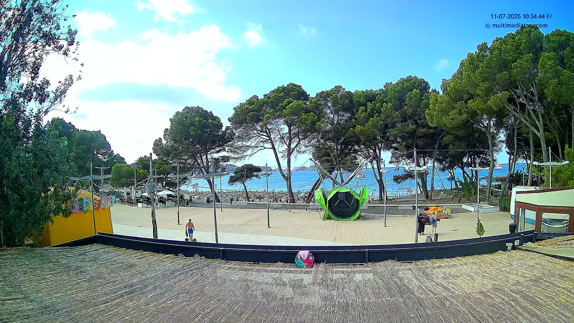 A sunny outdoor scene shows a paved plaza with a green attraction, several people, and tall pine trees overlooking a beach and calm blue ocean.