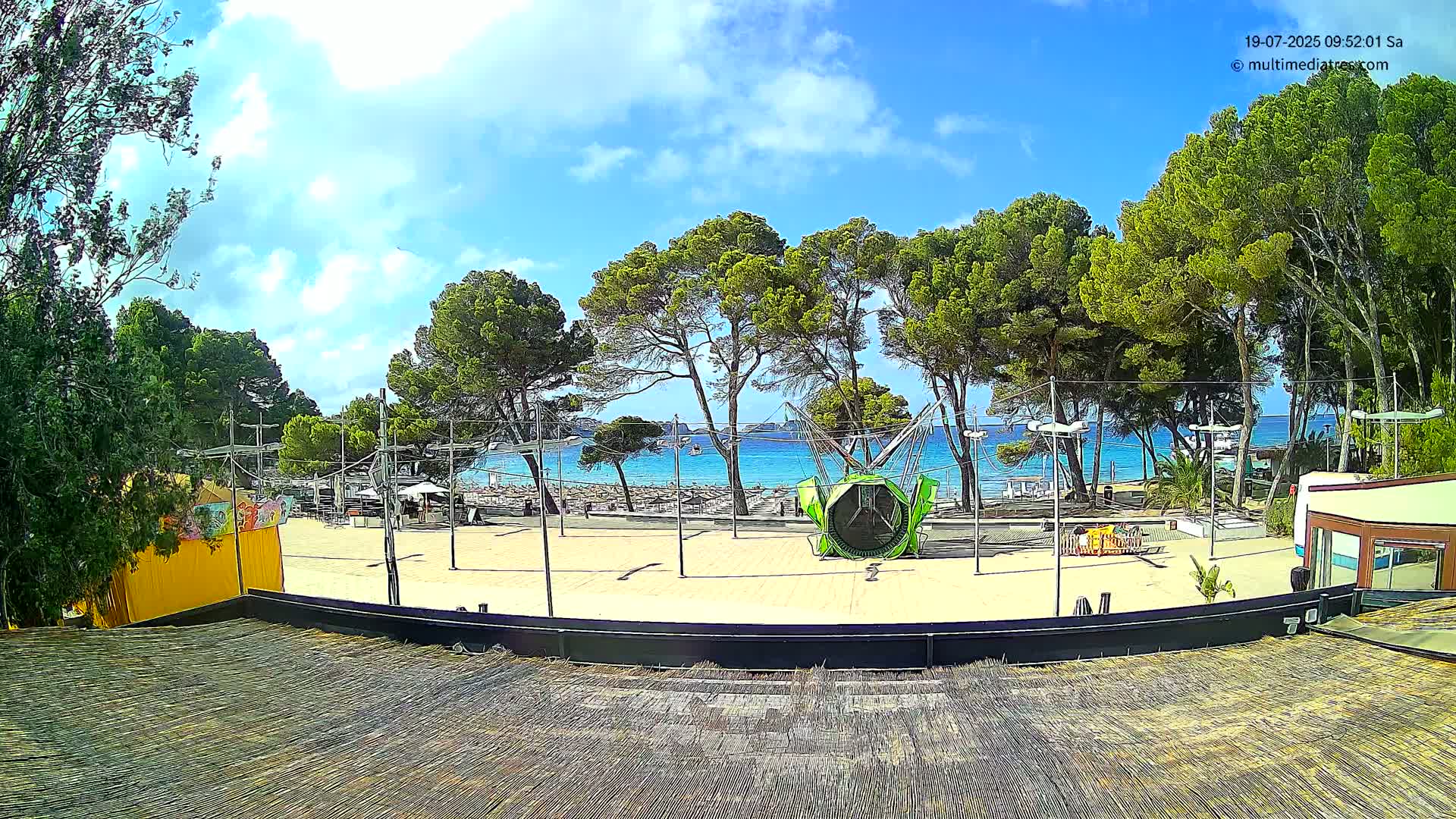 A sandy beachfront area with green trees, a playground ride, and a calm, blue ocean under a partly cloudy sky.