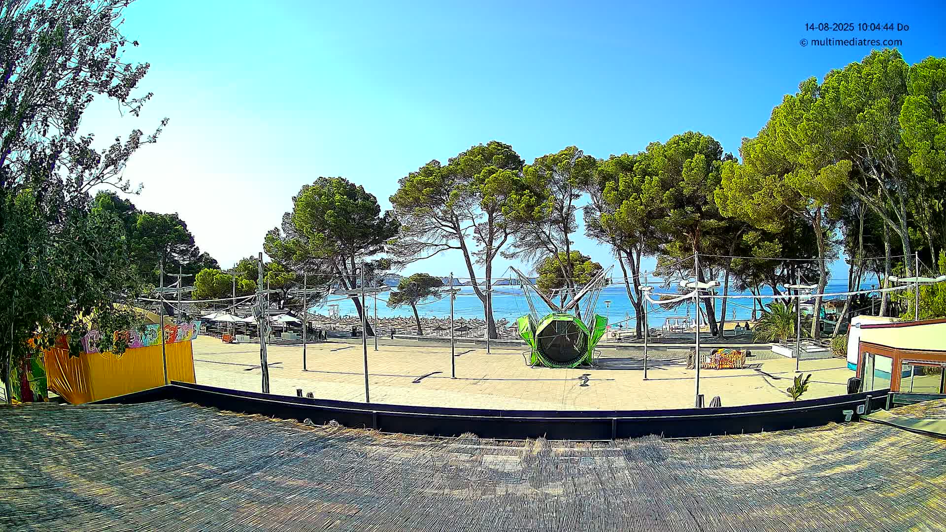 A sunny outdoor scene shows a paved plaza with a large, circular ride, bordered by trees and overlooking a beach with calm water.