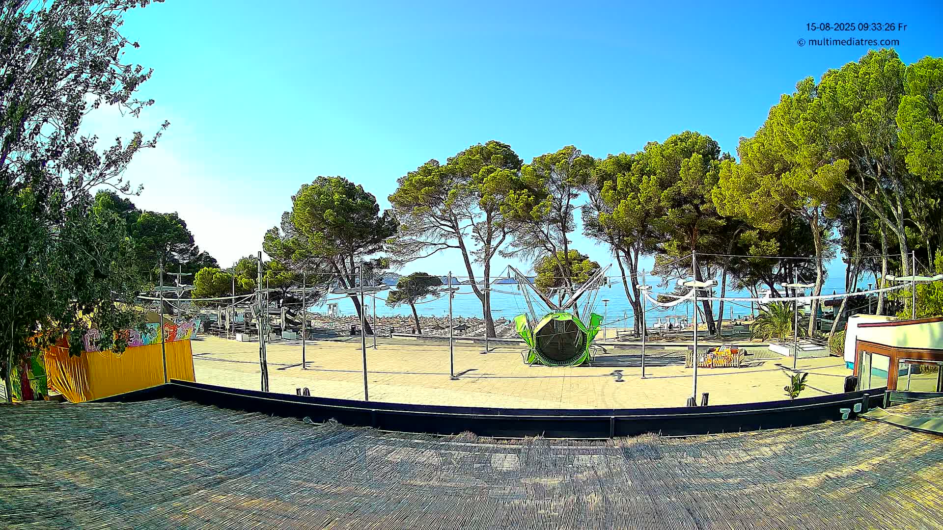 A sunny outdoor scene shows a paved area with a playground-like structure and a beach visible beyond a line of pine trees.