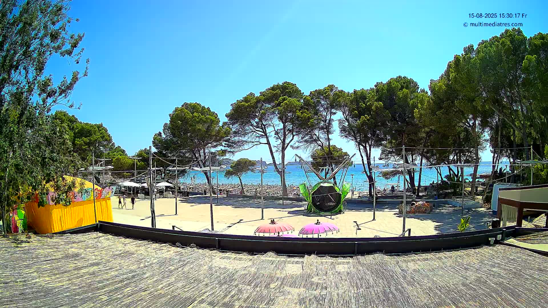 A sunny outdoor scene shows a paved area with a playground-like structure and a beach visible beyond a line of pine trees.