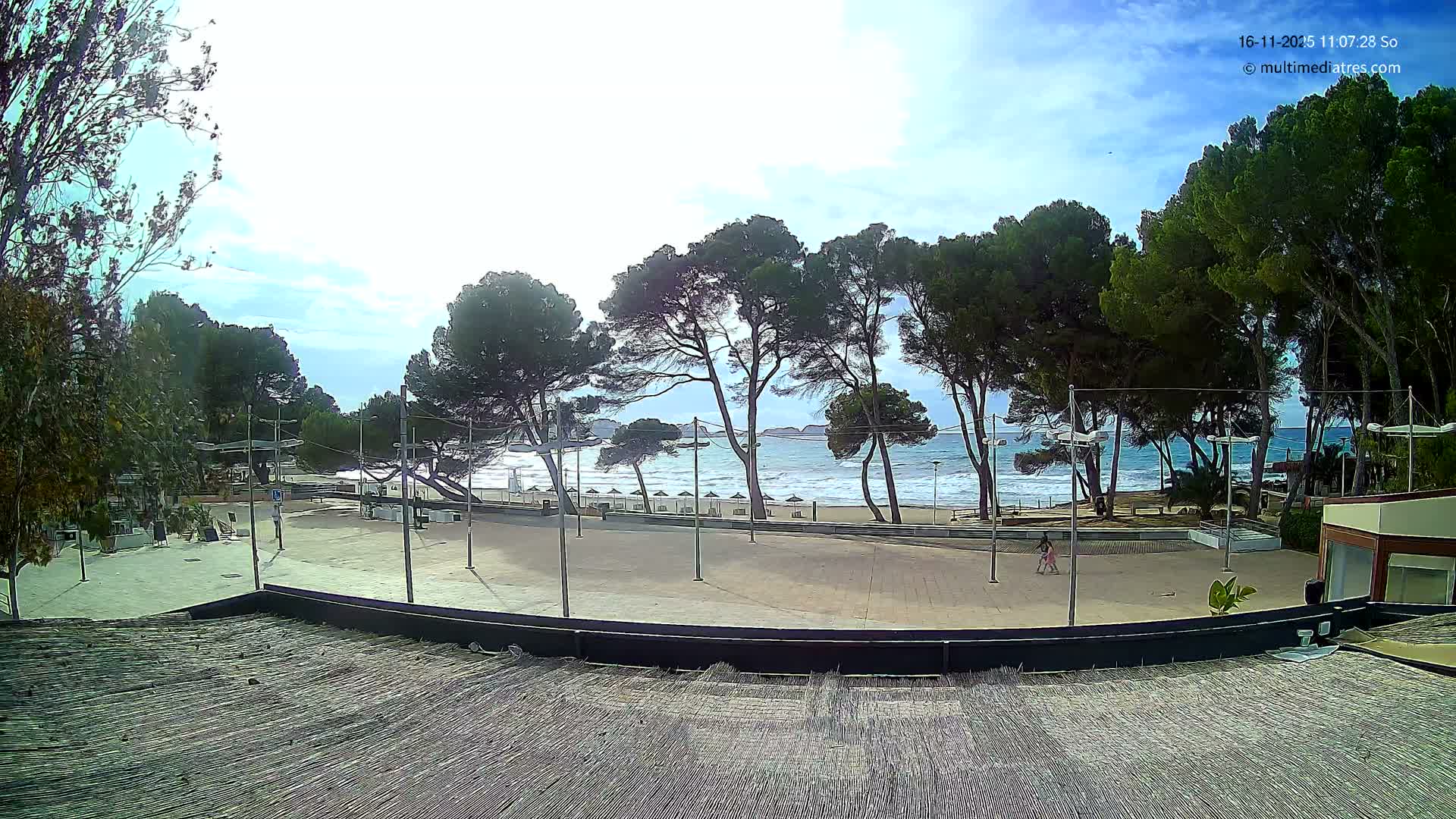 A sunny, partly cloudy day is depicted at a coastal scene with a sandy beach, crashing waves, and numerous pine trees, fronted by a paved promenade where a person walks, all viewed from above a textured roof or deck.