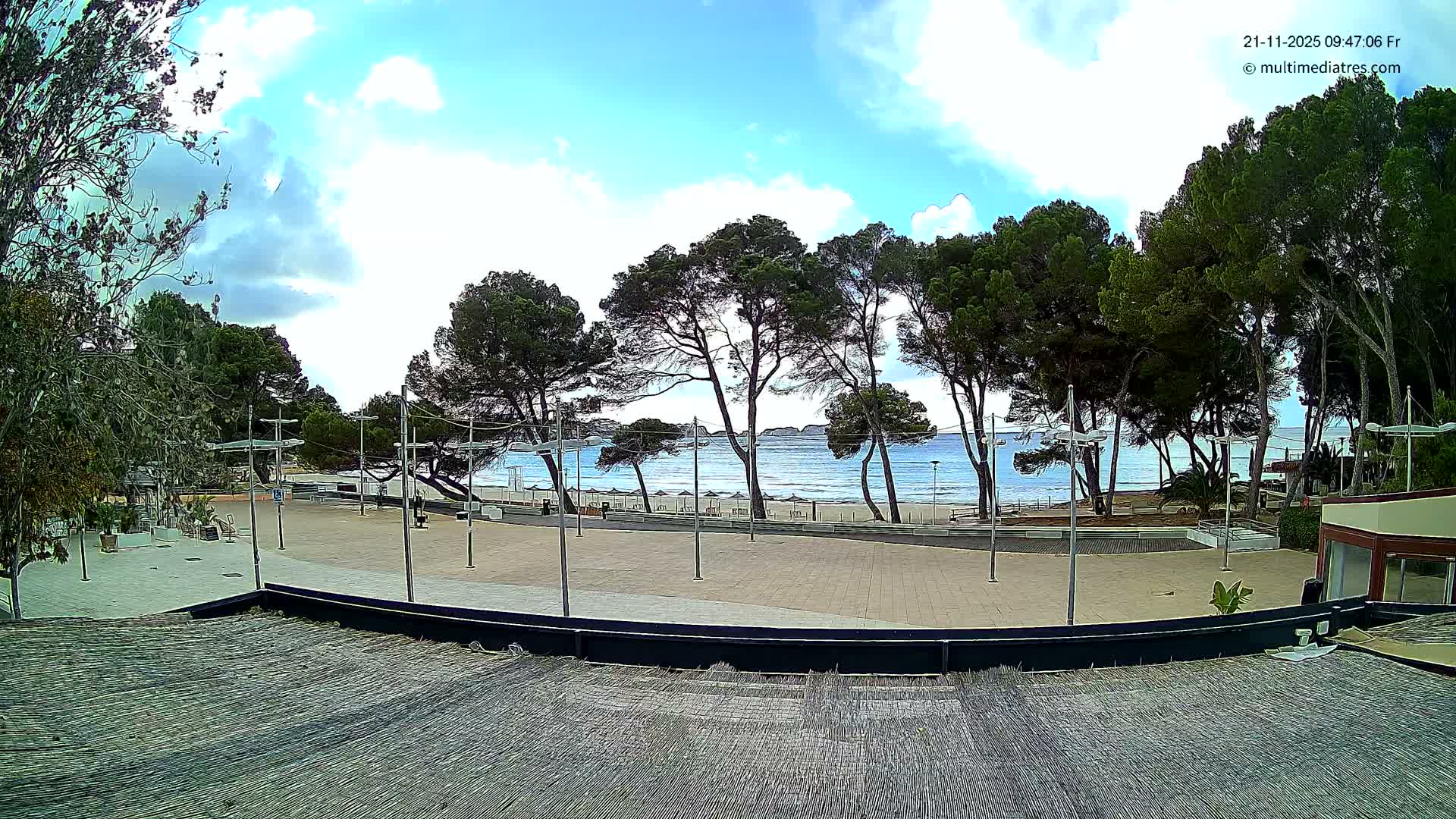 Under a partly cloudy sky, a sunny coastal view captures a wide paved area in the foreground, backed by a line of tall pine trees fronting a sandy beach with many sun loungers and a calm blue sea.