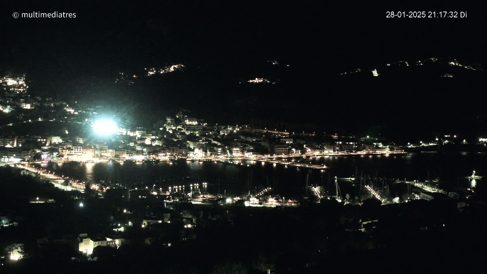 A nighttime view of a coastal town illuminated with lights, nestled between hills, overlooking a harbor filled with boats.