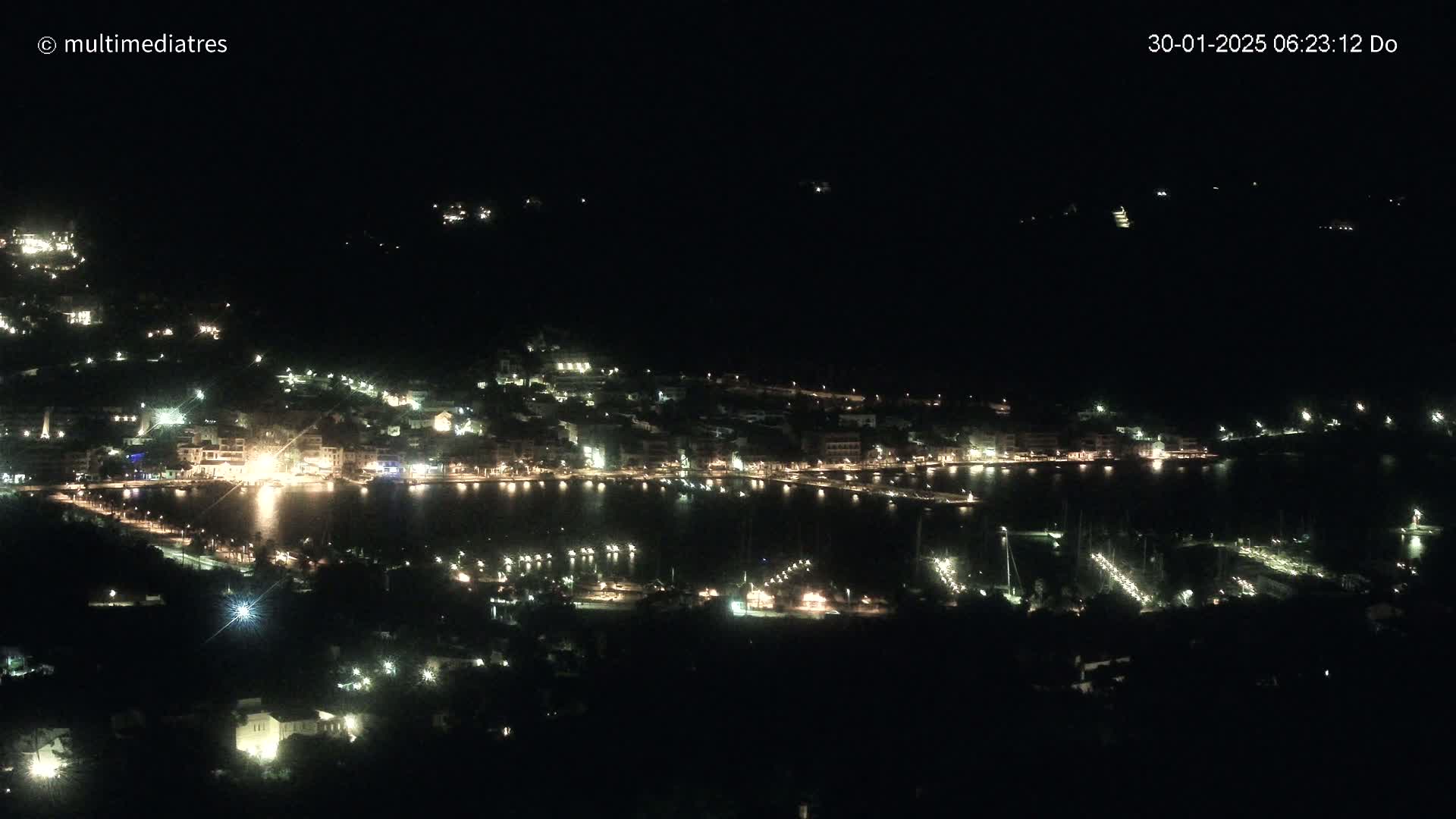 A nighttime, high-angle view of a coastal town illuminated by lights, with a dark body of water reflecting the lights.