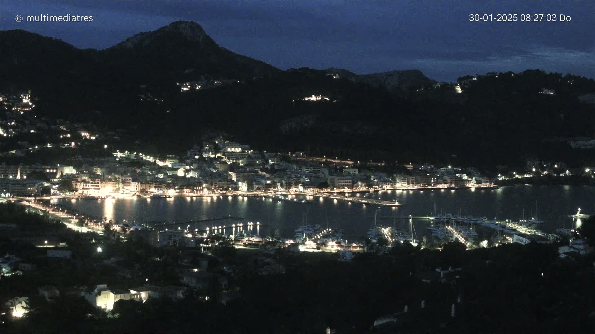A nighttime view of a harbor town nestled at the base of a dark mountain, illuminated by many lights.