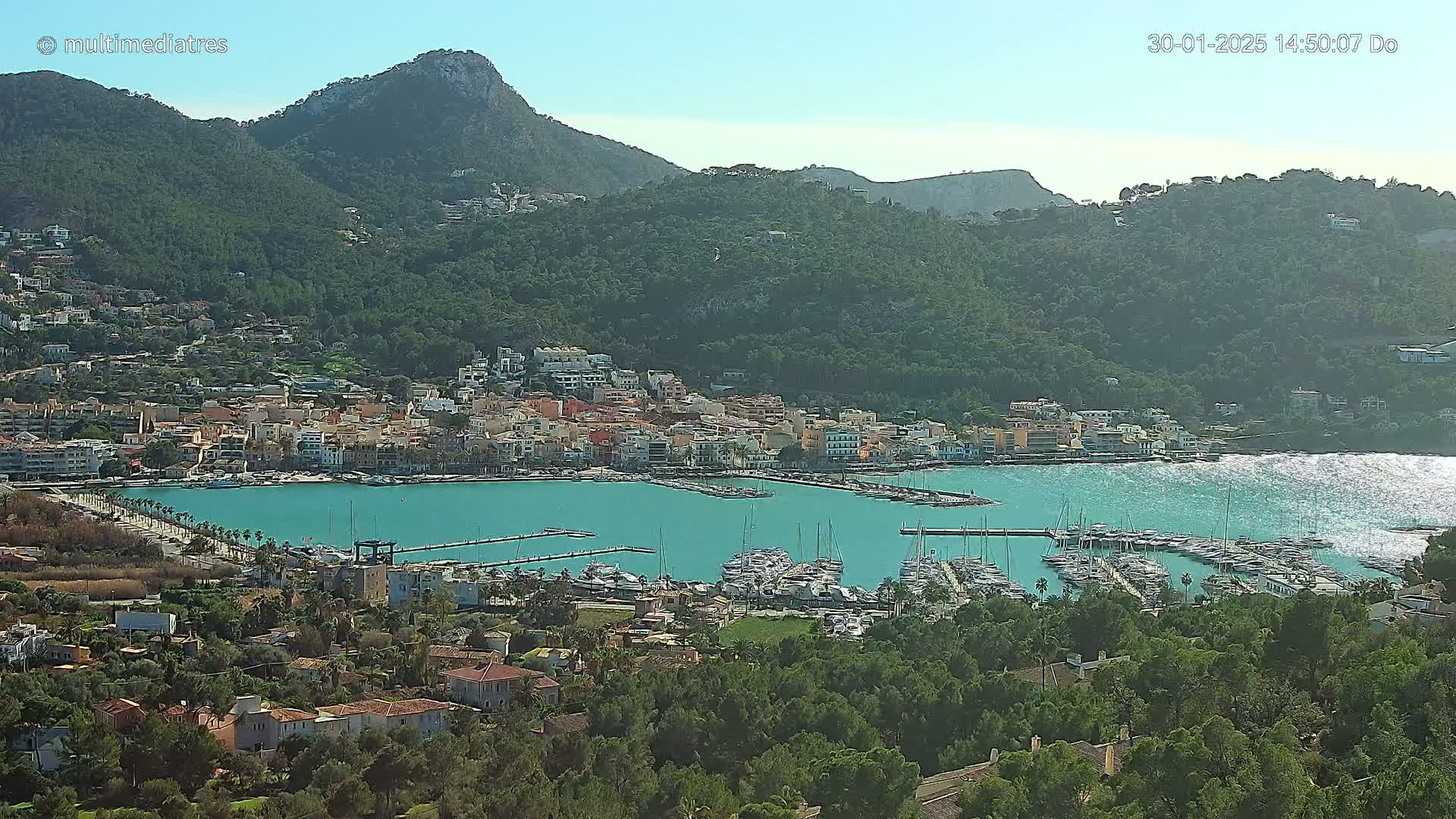 A coastal town nestled against a mountainside, with a harbor filled with sailboats under a clear, sunny sky.