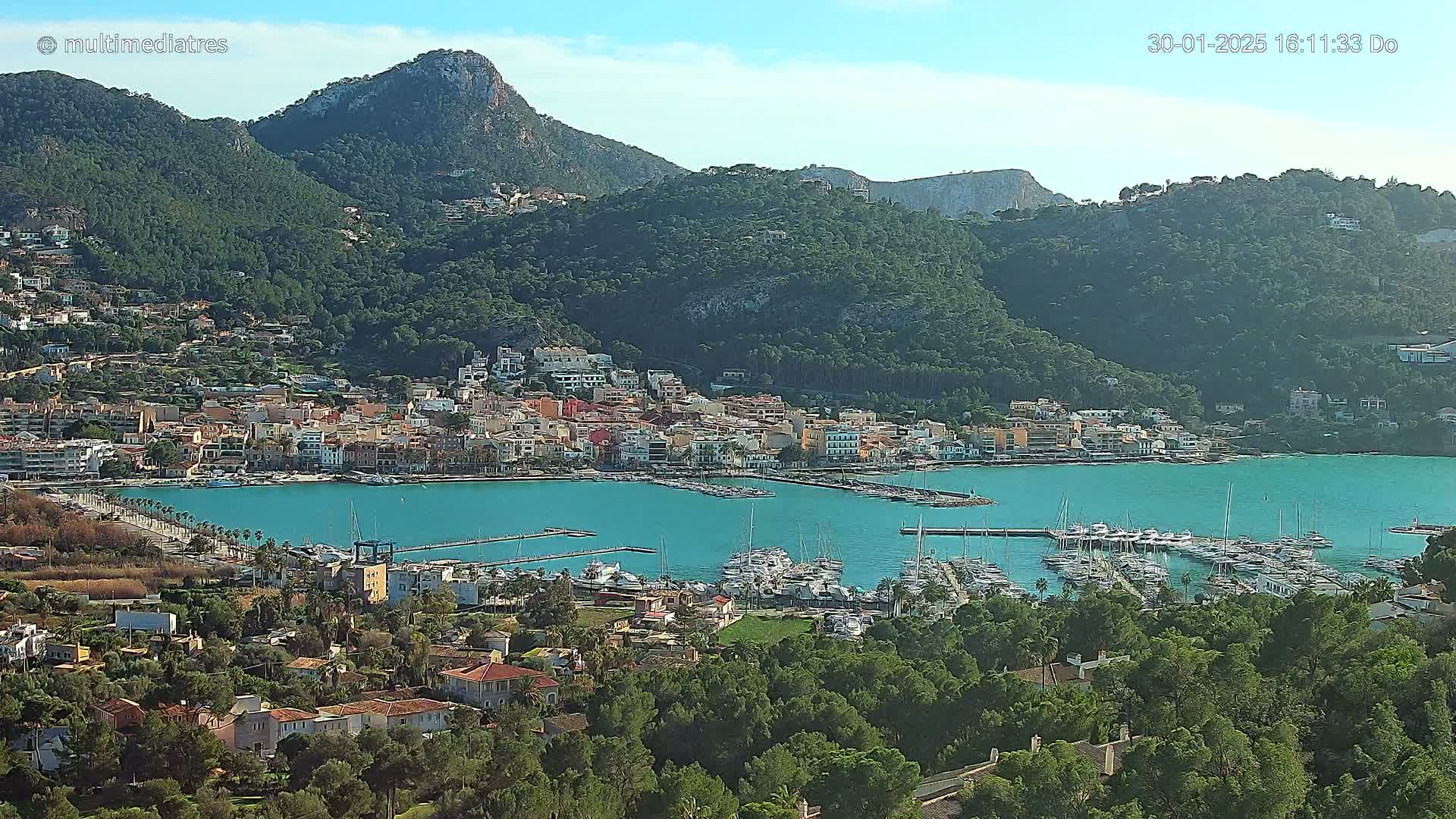 A coastal town nestled between green hills and a turquoise bay filled with numerous boats, under a clear sunny sky.
