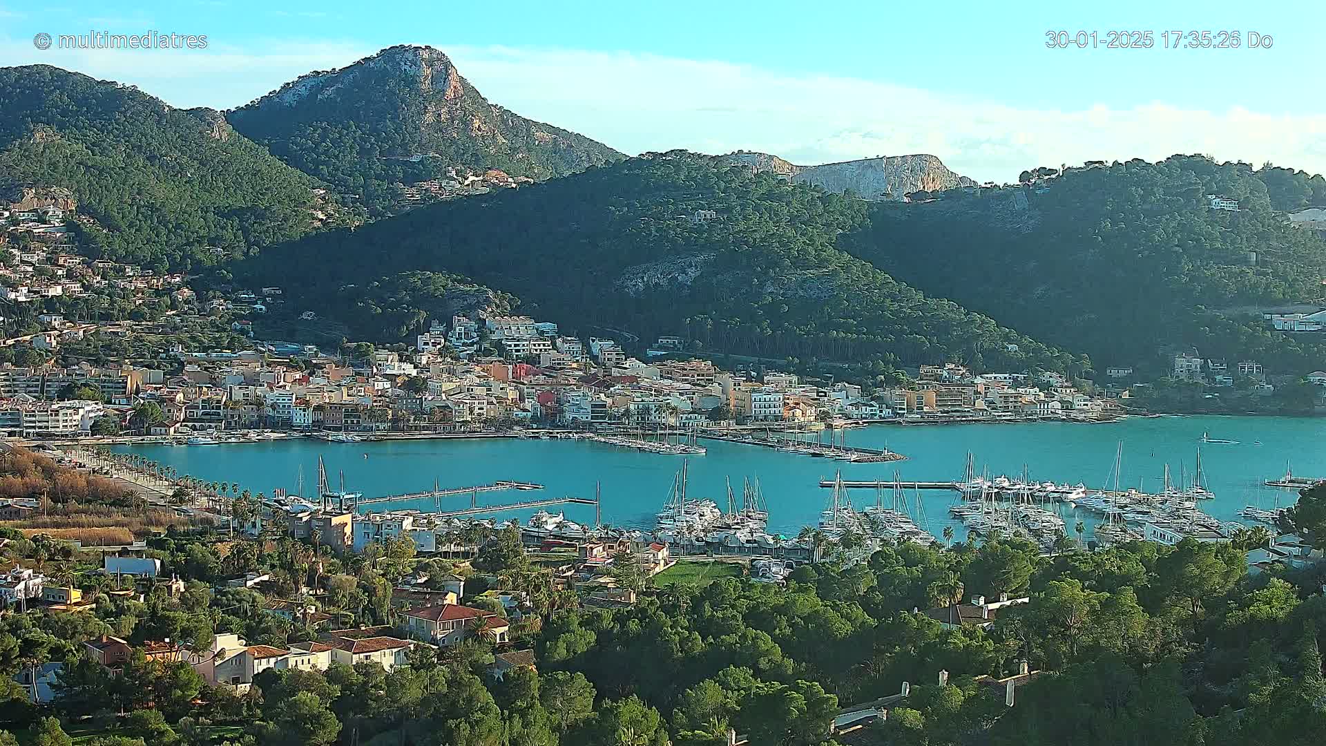 A coastal town nestled between green hills and a clear blue harbor filled with sailboats, under a bright, sunny sky.