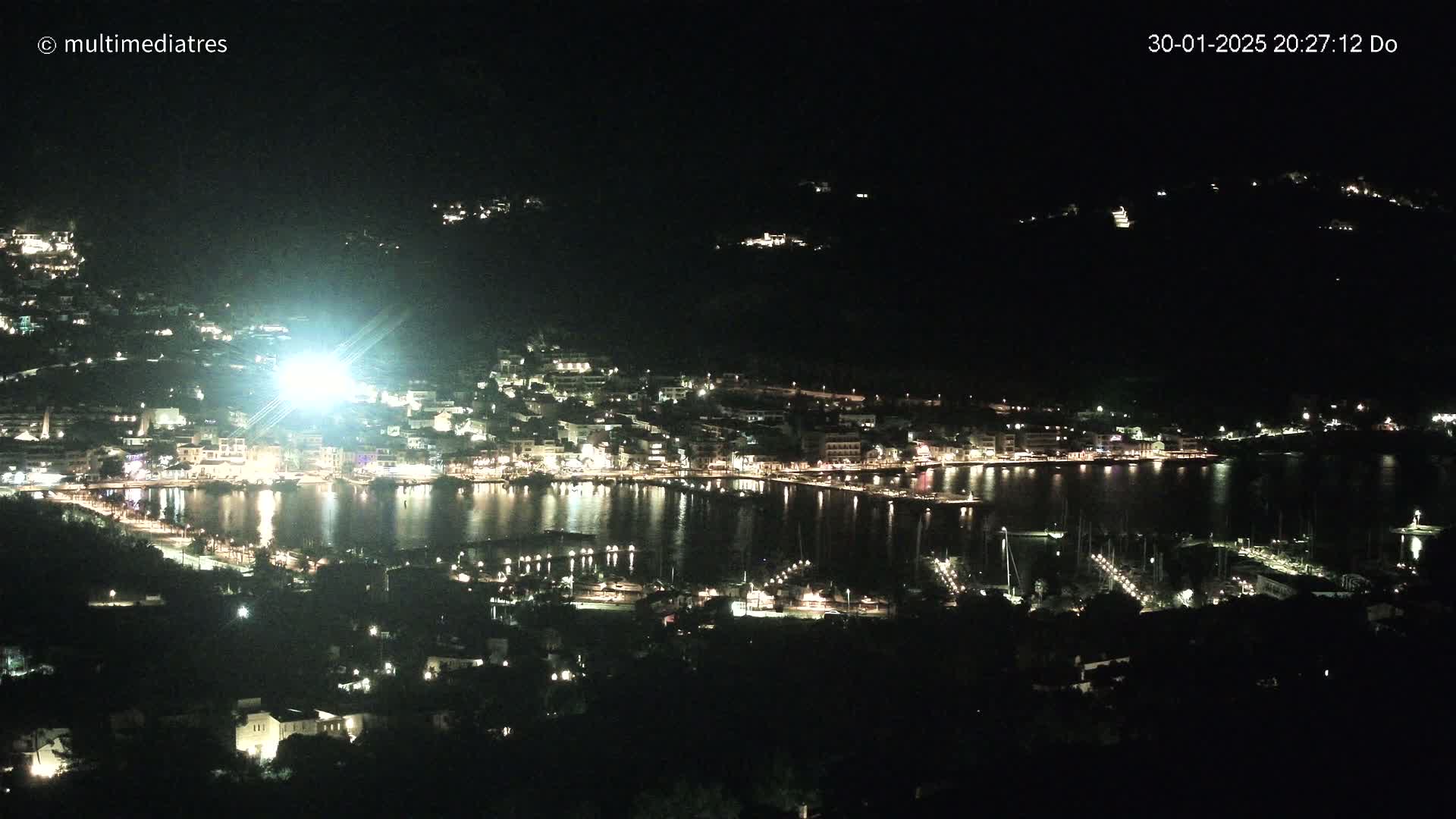 A nighttime view of a brightly lit coastal town and harbor, nestled among dark hills.