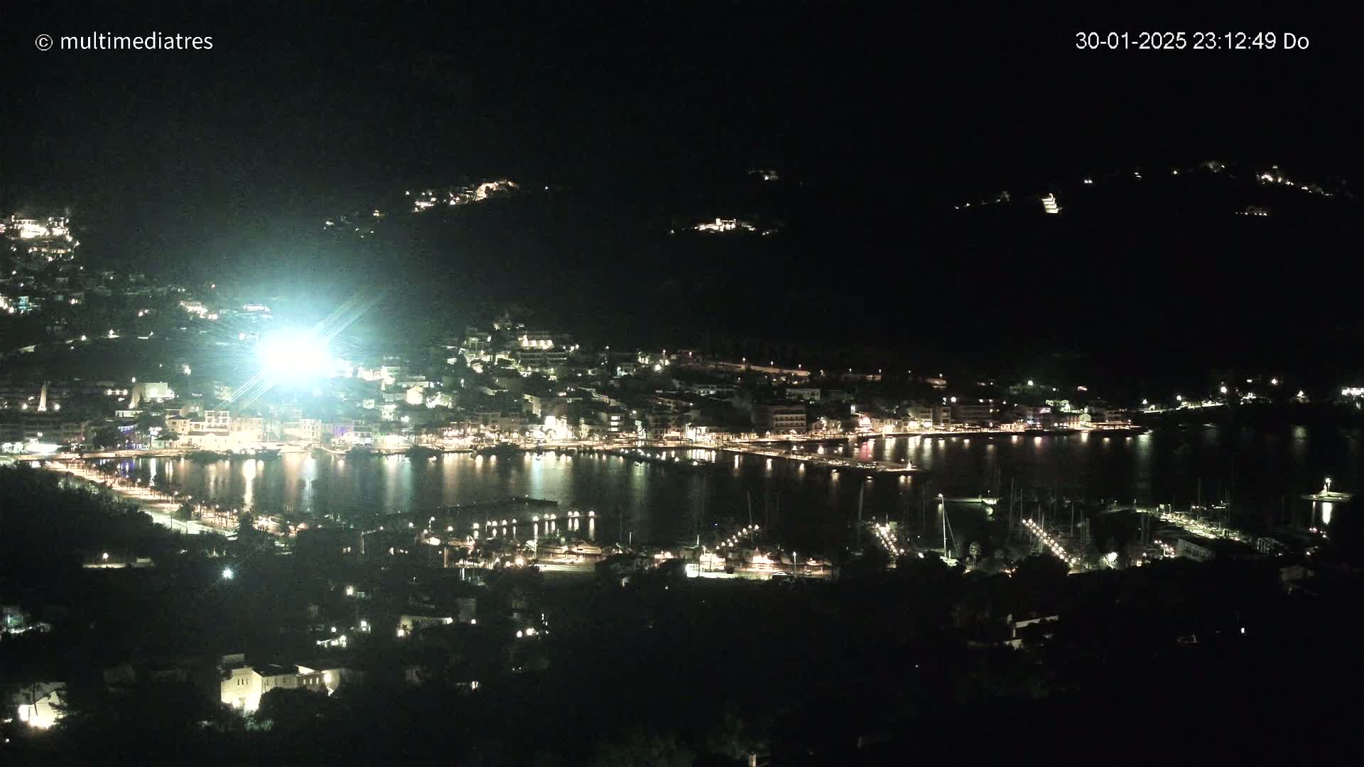 A brightly lit coastal town at night is nestled in a dark, hilly landscape, with a harbor filled with boats.