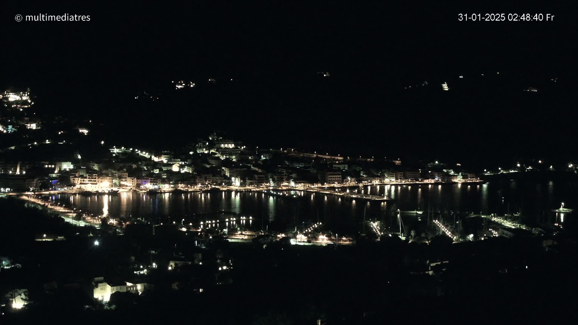 A nighttime view of a coastal town with its lights illuminating the harbor and surrounding hills under a clear, dark sky.