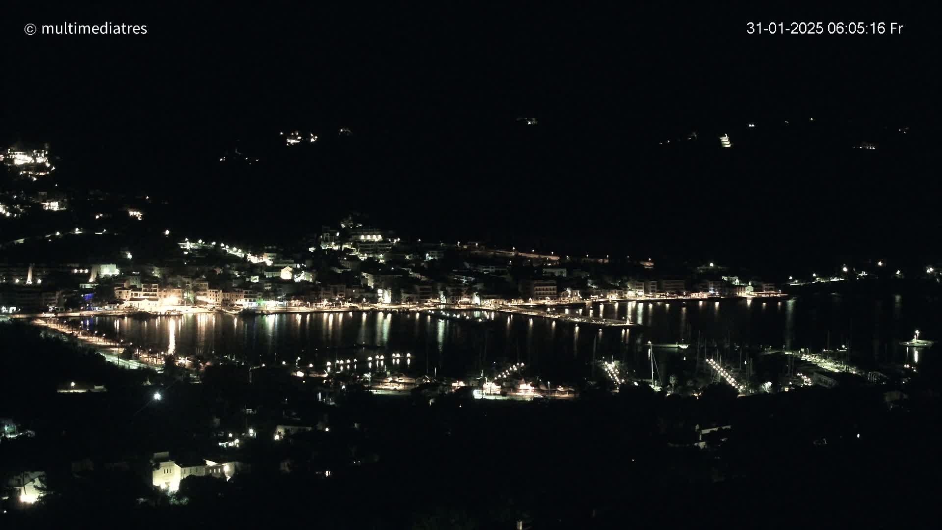 A nighttime, high-angle view of a harbor town illuminated by lights, nestled along a dark shoreline.