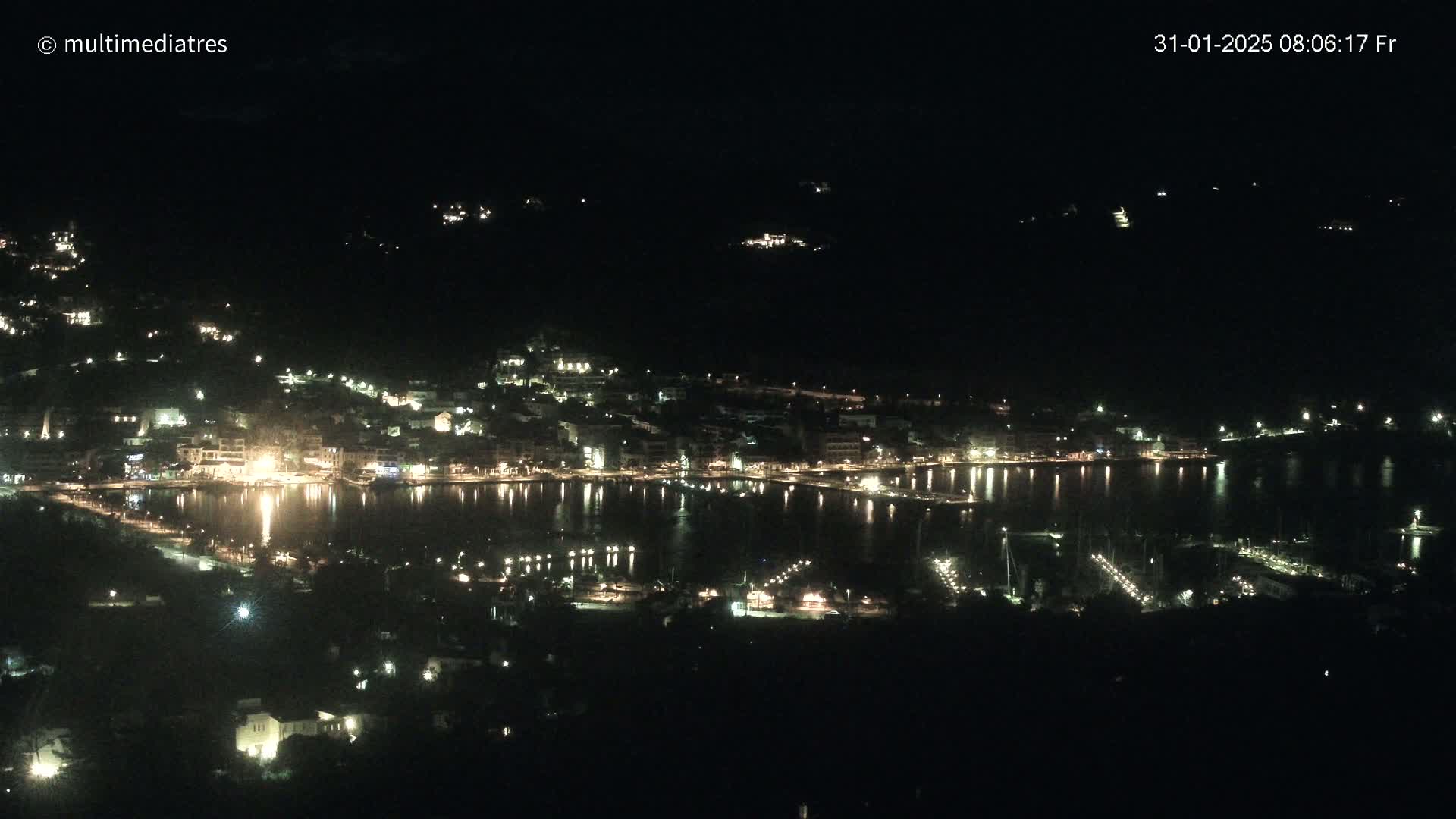 A nighttime, high-angle view shows a coastal town with its lights reflected in a calm body of water.