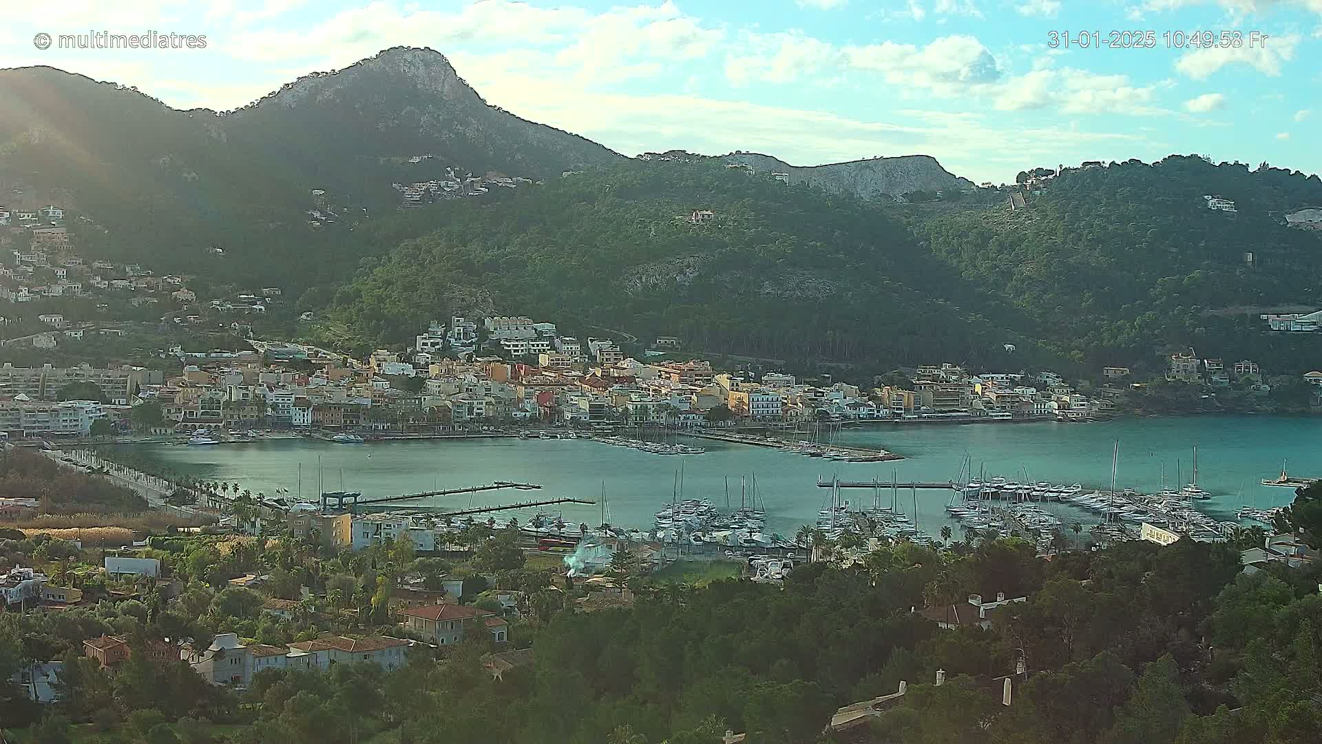 A coastal town nestled in a mountainous region, with a marina filled with boats under a partly cloudy sky.