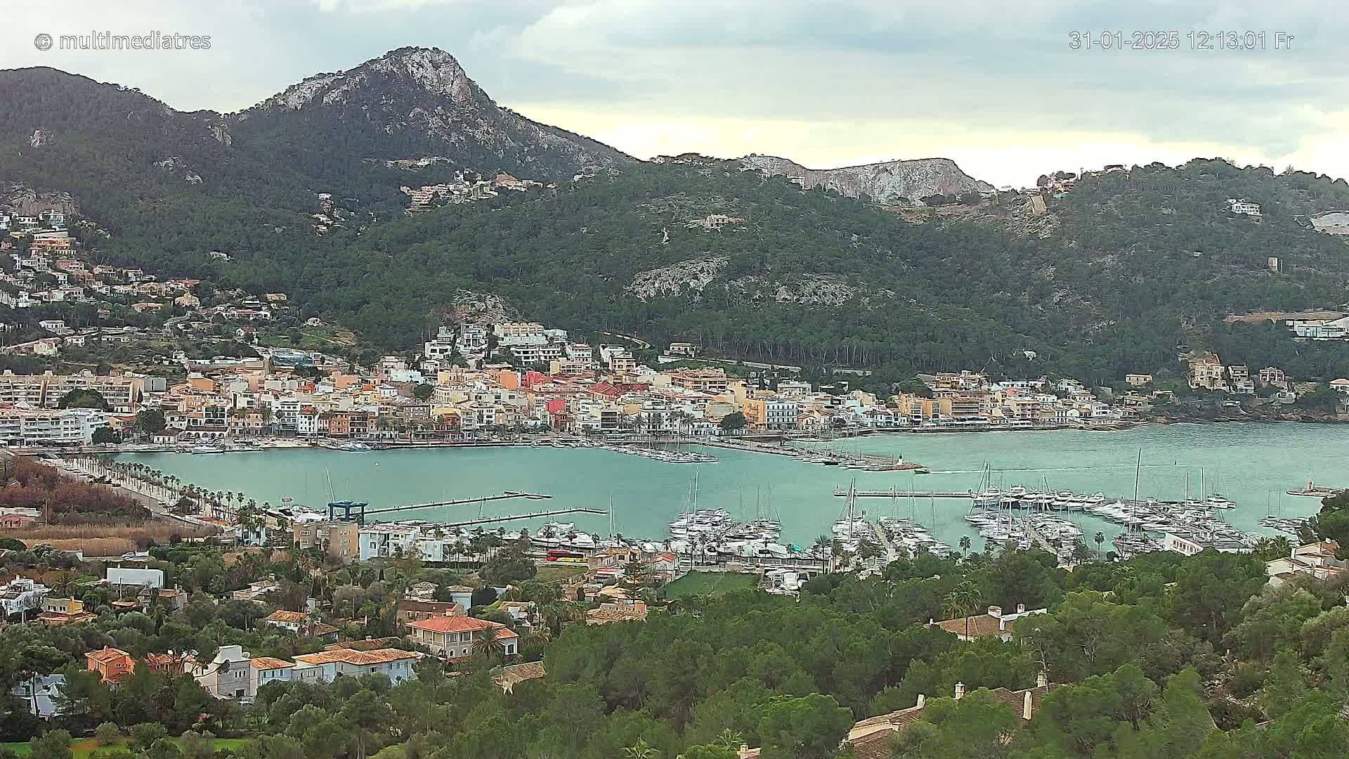 A picturesque coastal town nestled at the base of a mountain range under an overcast sky, featuring a marina filled with numerous boats.