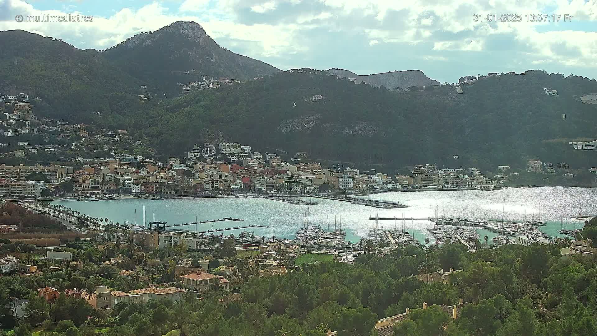 A coastal town nestled in a mountainous region with a marina filled with boats under partly cloudy skies.