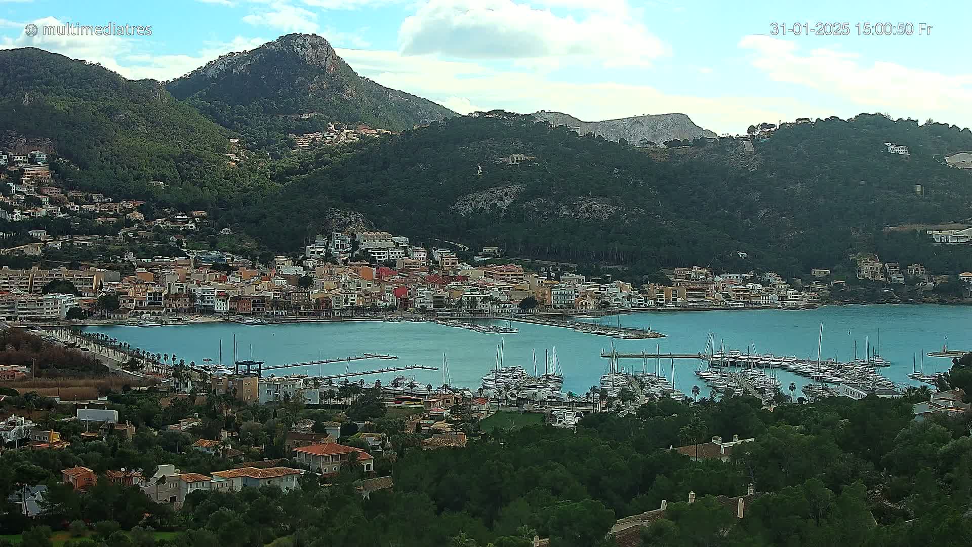 A coastal town nestled between hills under a partly cloudy sky, features a marina filled with boats.