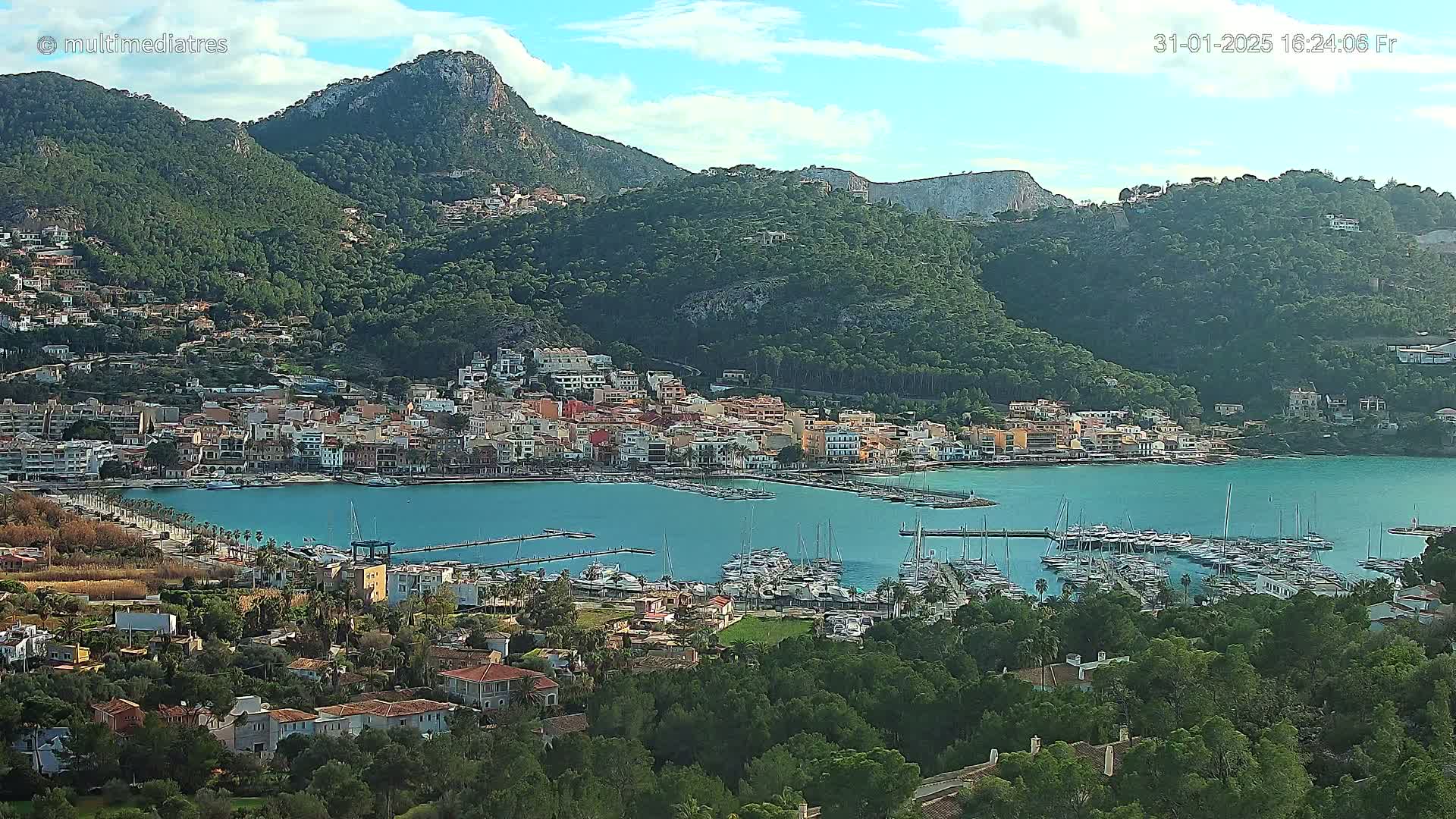A coastal town nestled between green hills and a calm, blue bay filled with numerous boats under a mostly clear sky.