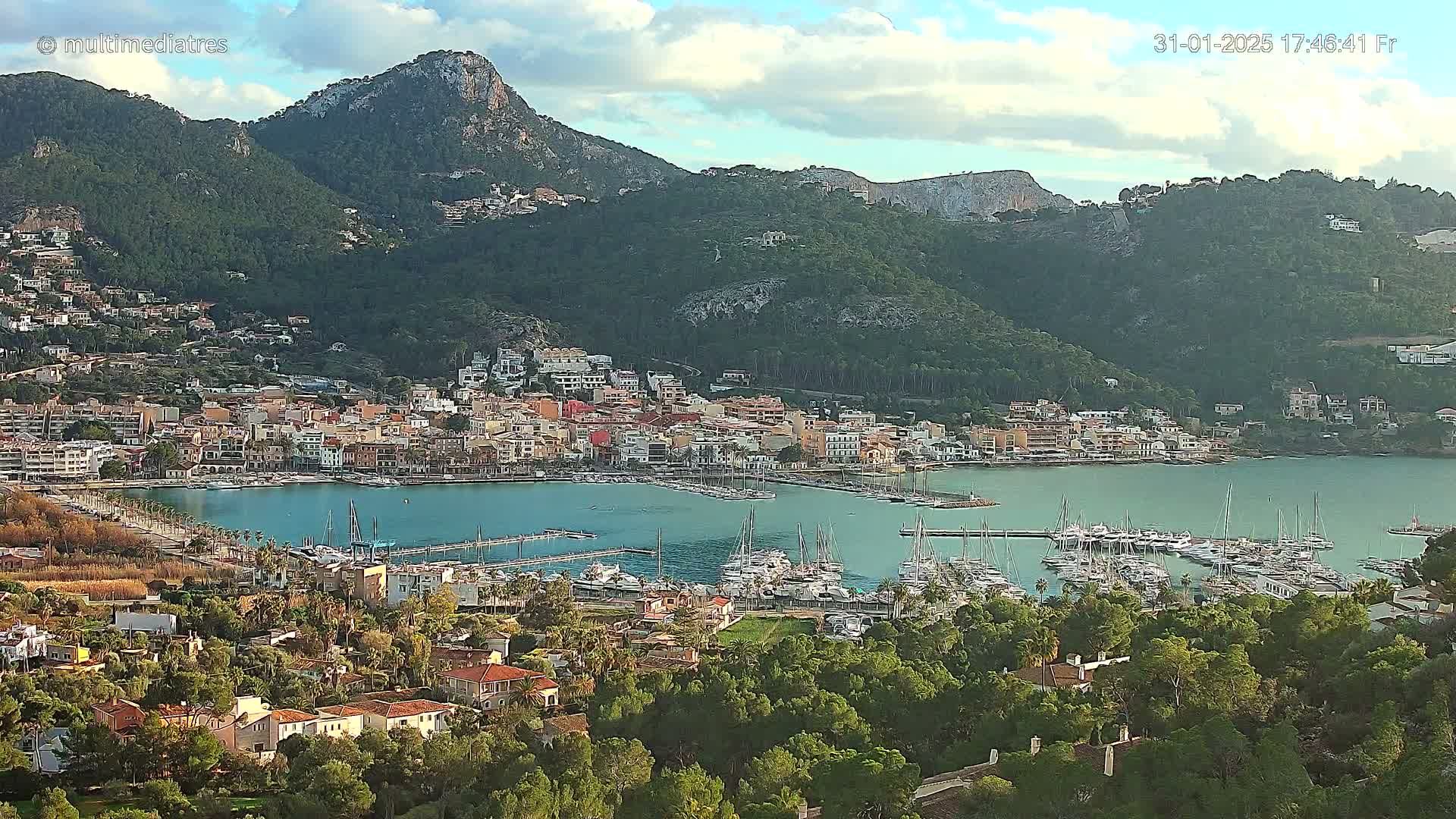 A coastal town with a marina filled with sailboats is nestled at the base of a mountain range under partly cloudy skies.