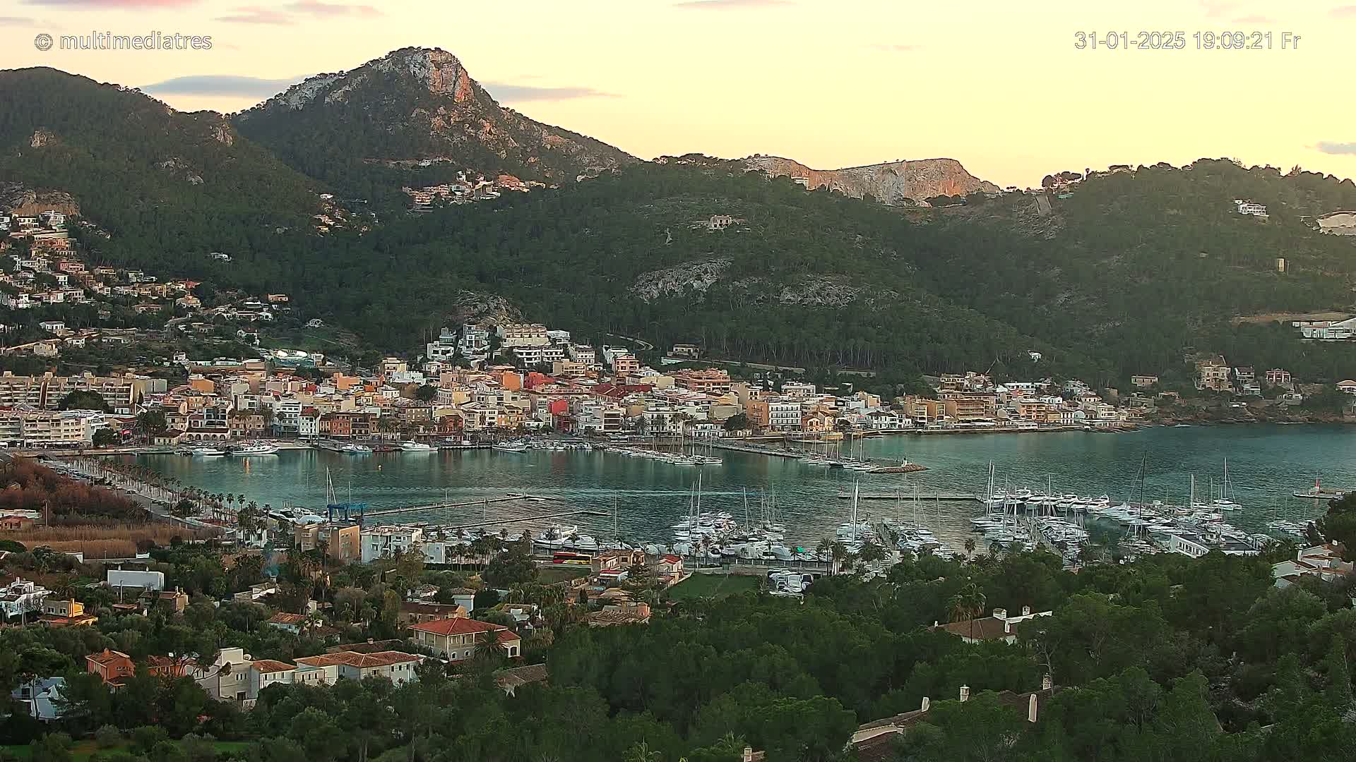 A coastal town nestled between mountains, with a calm harbor filled with numerous boats, under a partly cloudy sunset sky.