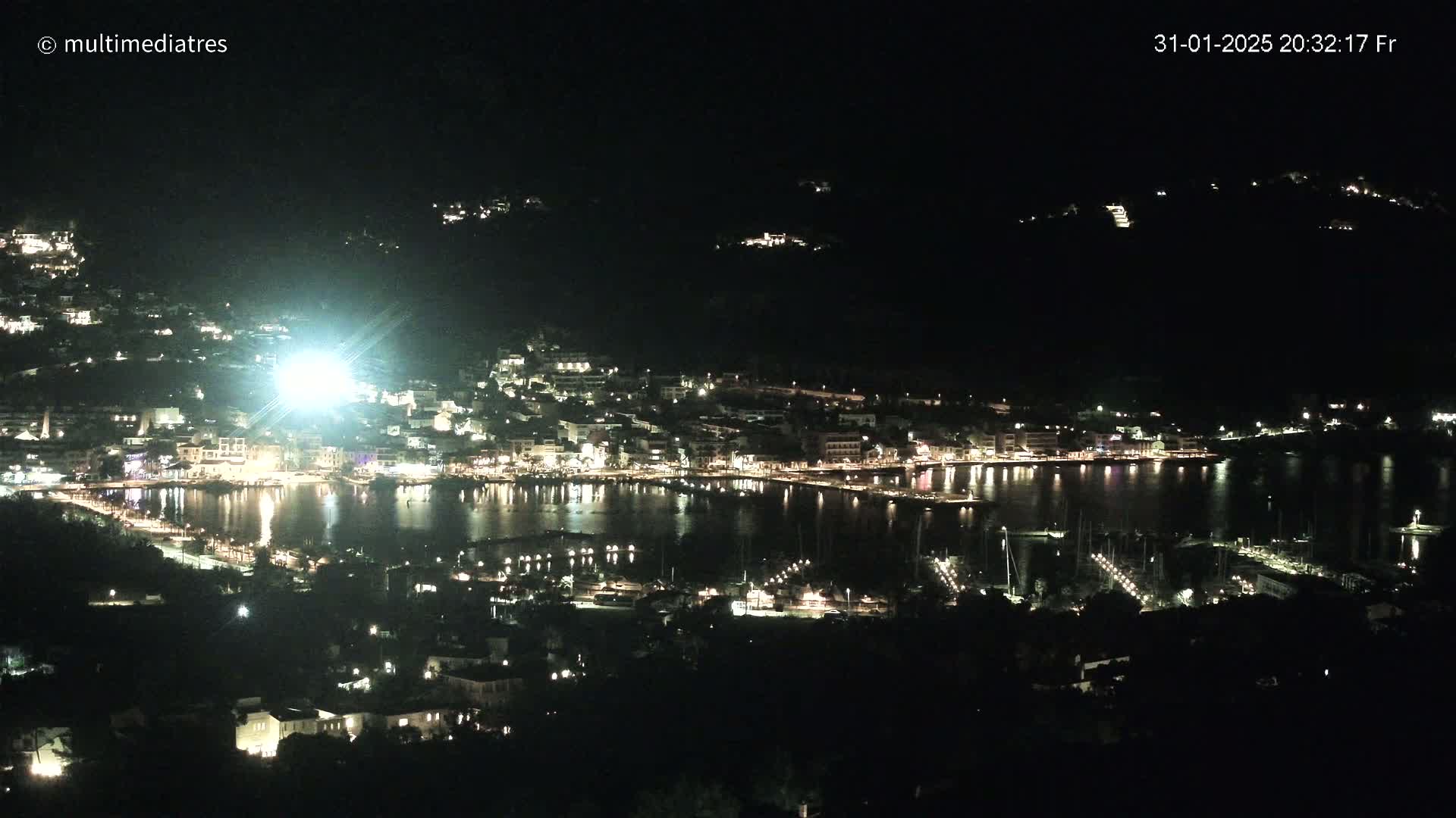 A nighttime, aerial view of a harbor town nestled among dark hills, illuminated by the lights of buildings and boats.