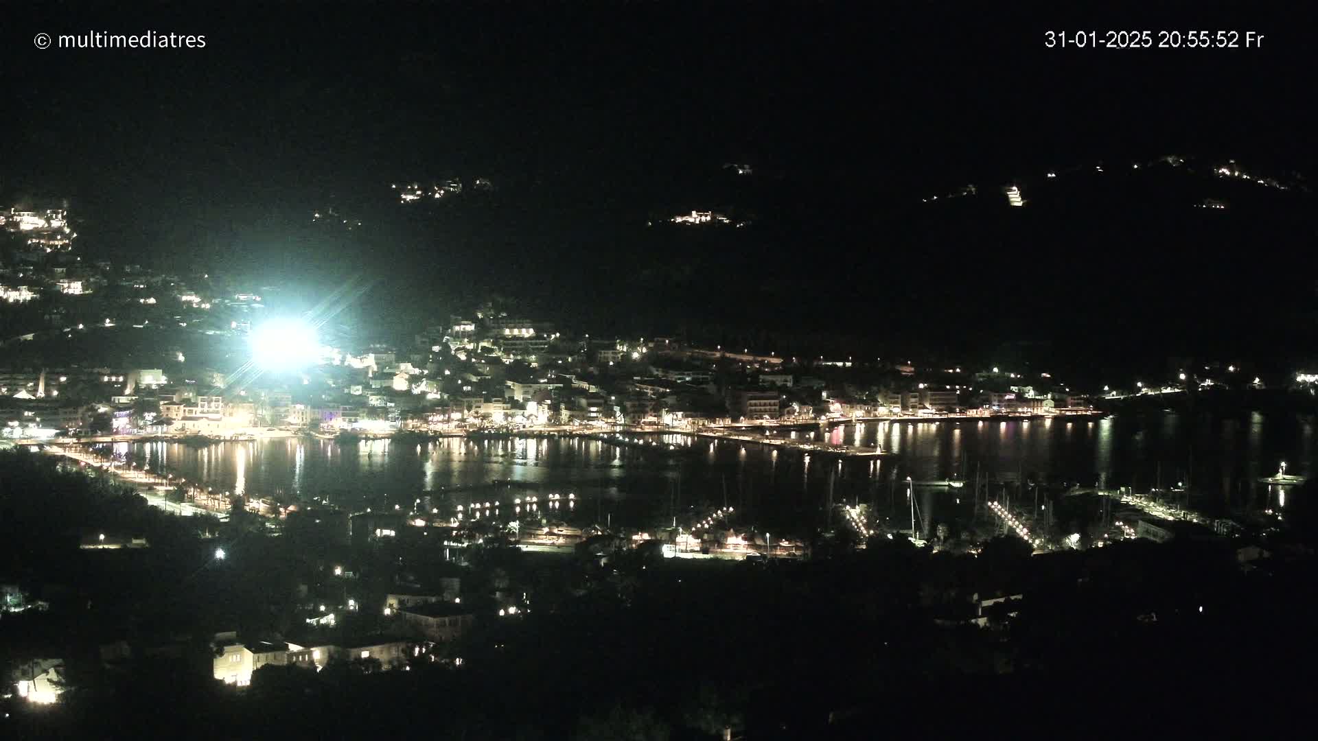 A nighttime, aerial view of a brightly lit town situated on a harbor, with boats docked and surrounding hills shrouded in darkness.