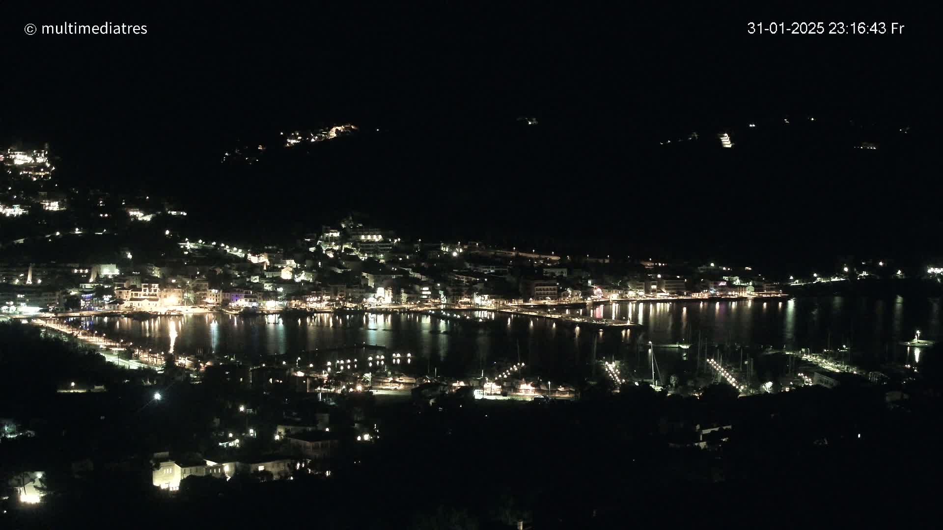 A nighttime view of a coastal town illuminated by lights, reflecting on calm water, under a clear, dark sky.