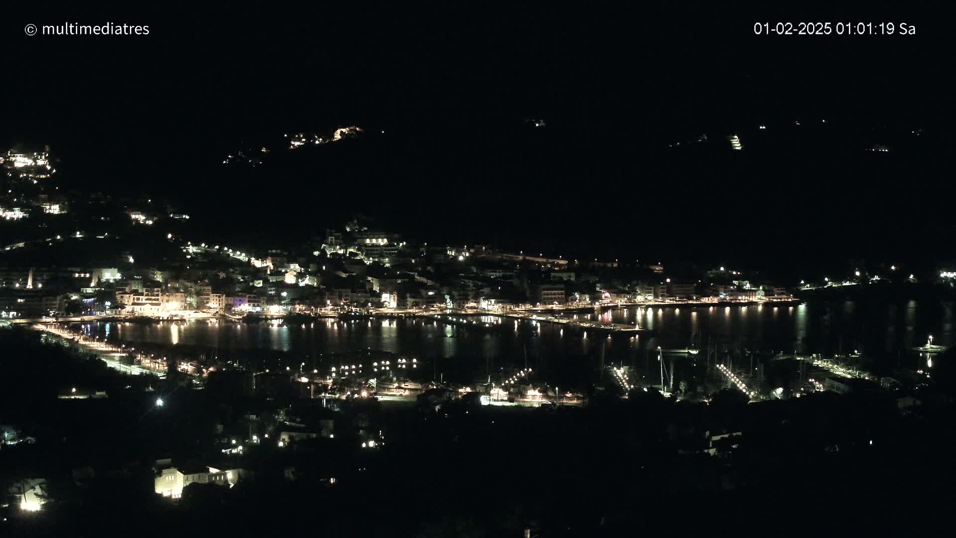 A nighttime view of a coastal town illuminated by lights, nestled along a dark body of water under a clear, dark sky.