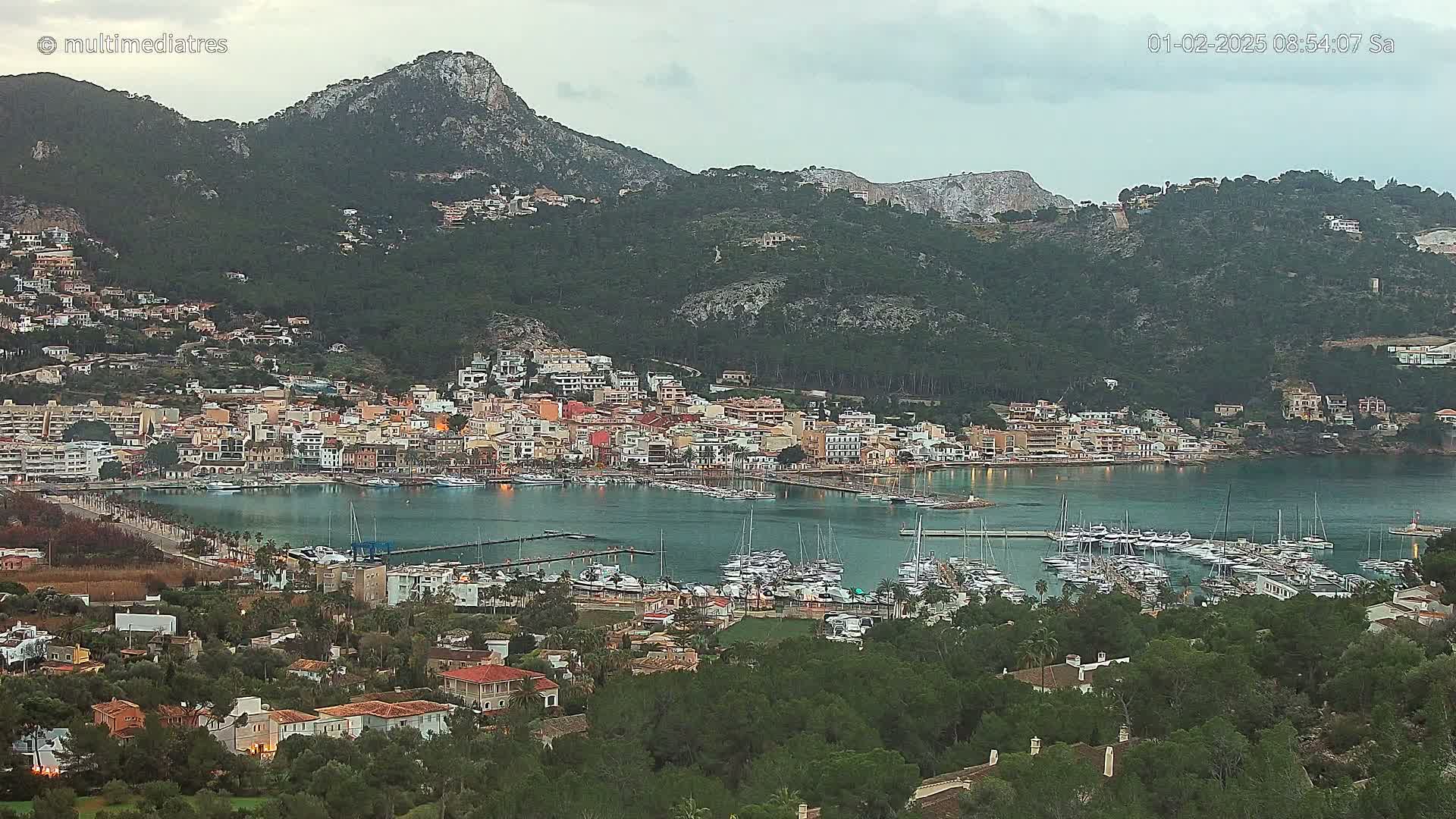 A coastal town nestled against a mountainside, with a calm harbor filled with sailboats under an overcast sky.