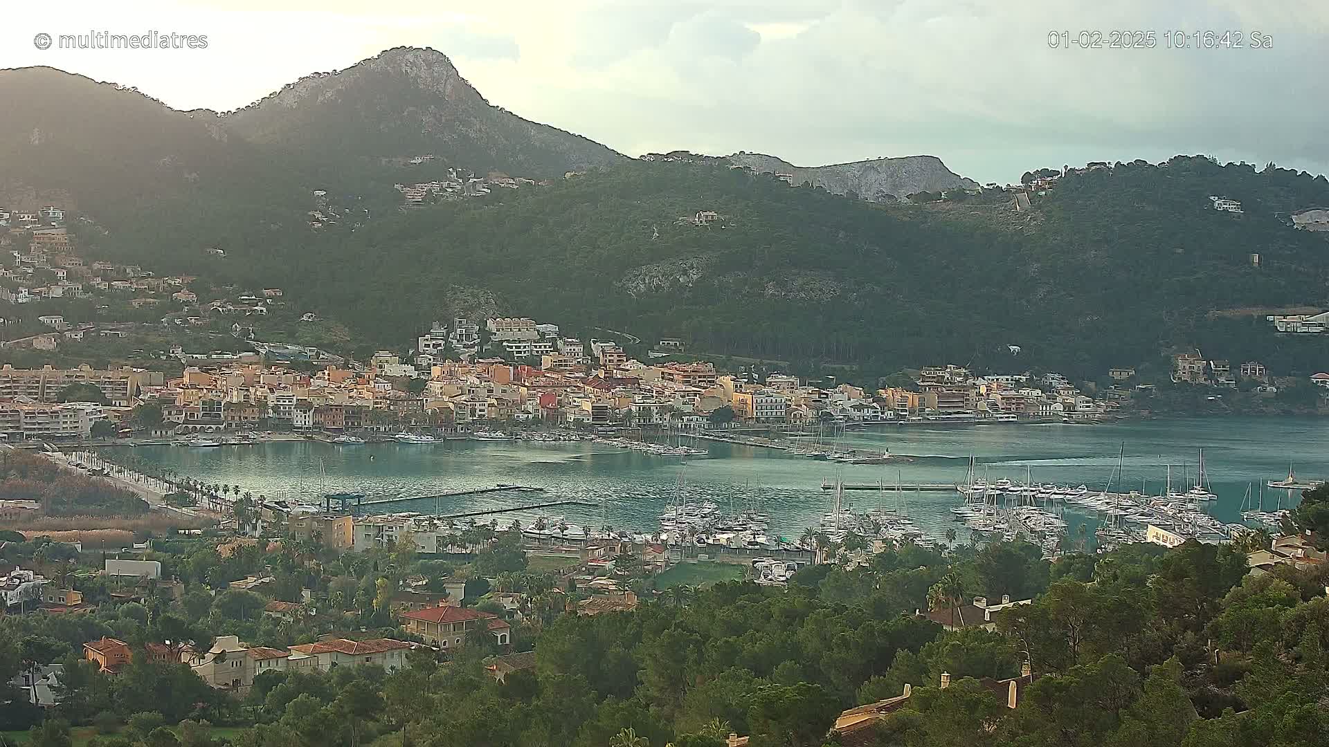 A coastal town with a marina full of boats sits nestled at the base of a large, green mountain range under an overcast sky.